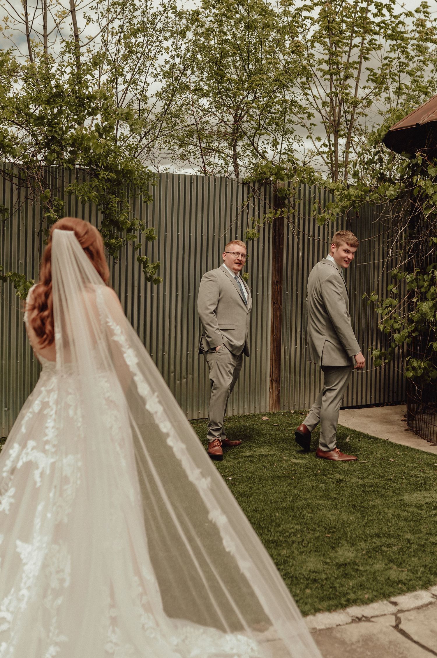 Two people in gray suits stand in a garden while a long white wedding veil flows in the foreground.