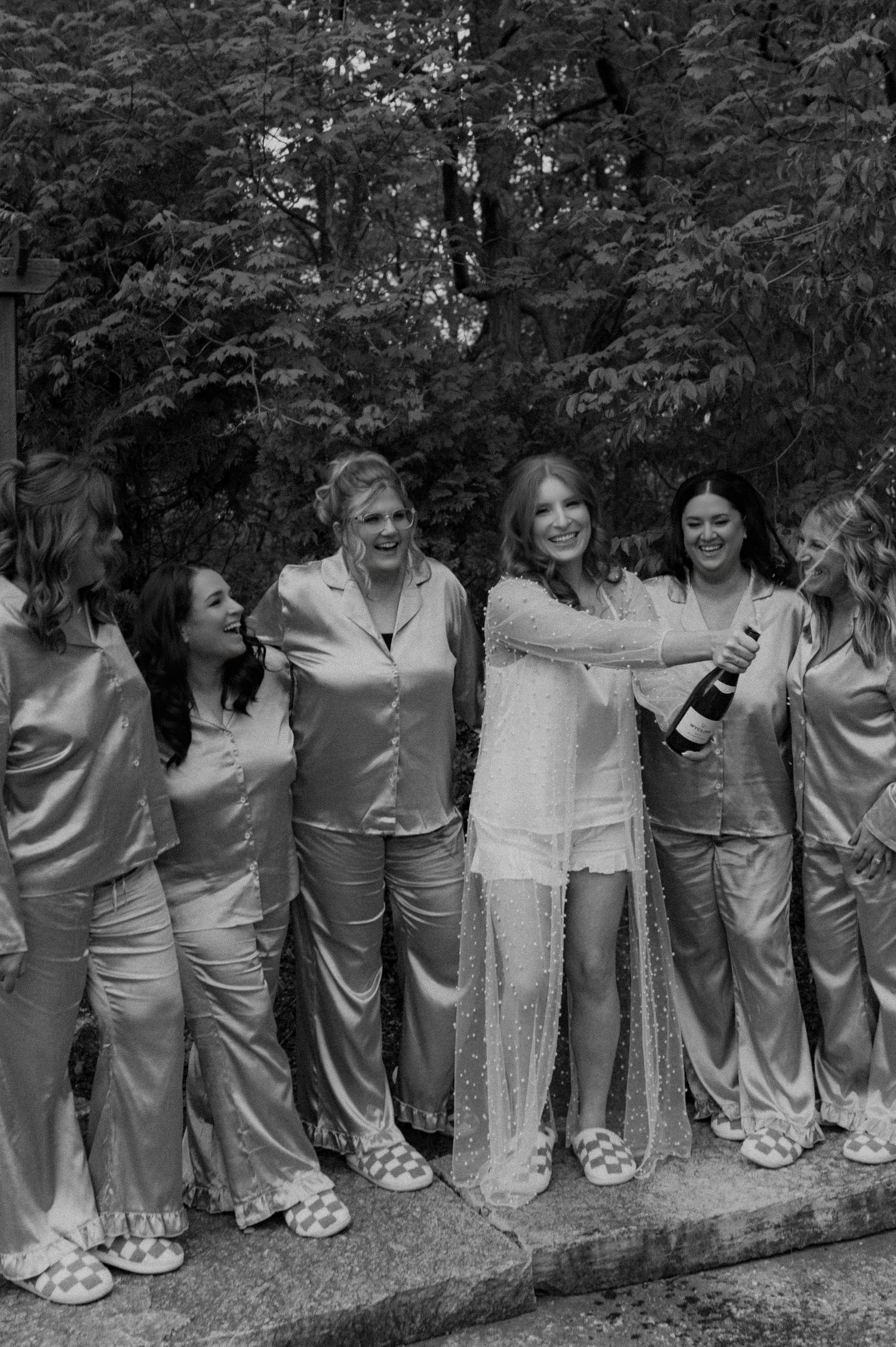 A group in matching silver dresses and white shoes stands in a row along a stone wall outdoors.