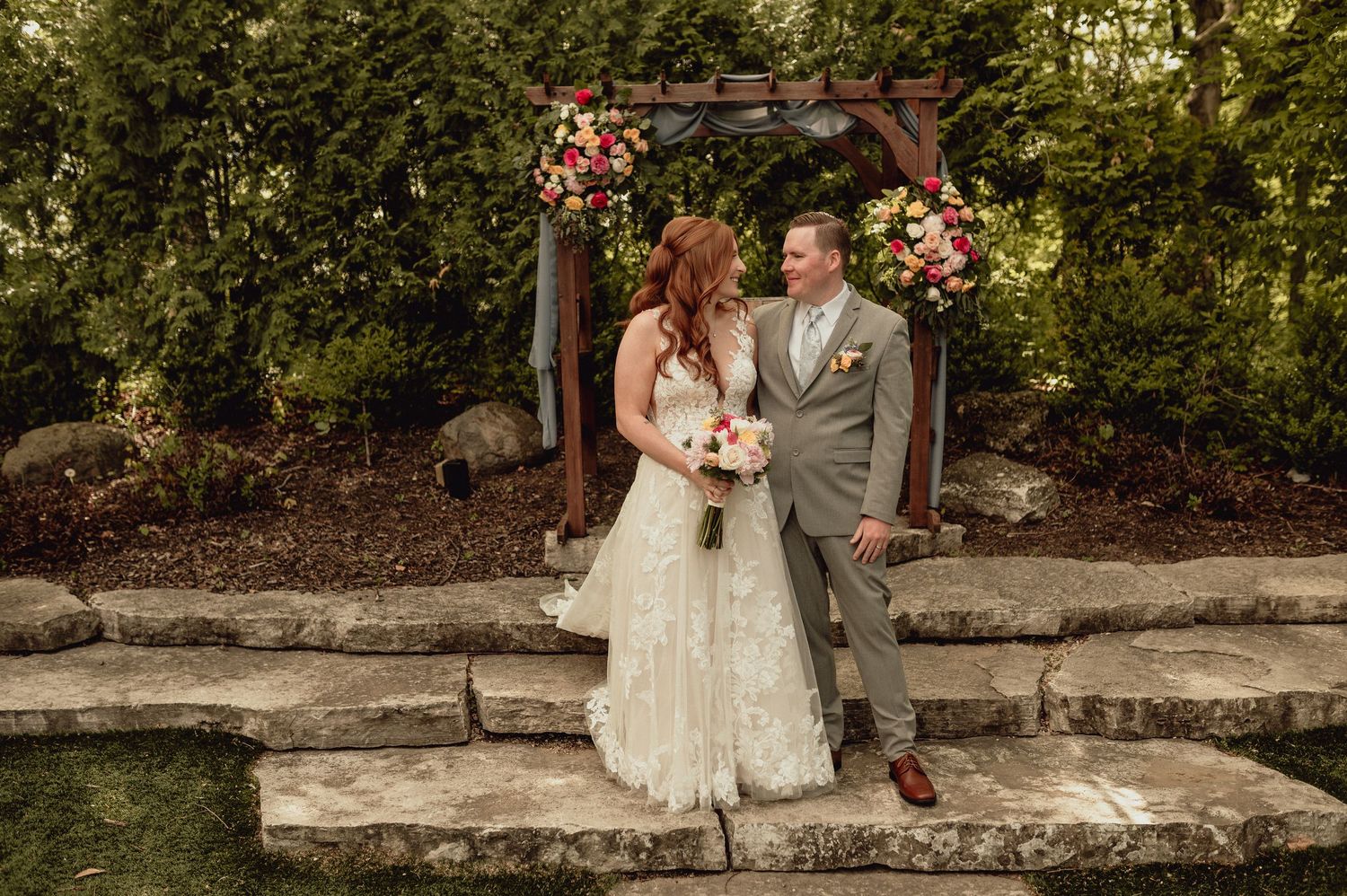 A couple in wedding attire poses for romantic photos on stone steps surrounded by lush greenery and gardens.