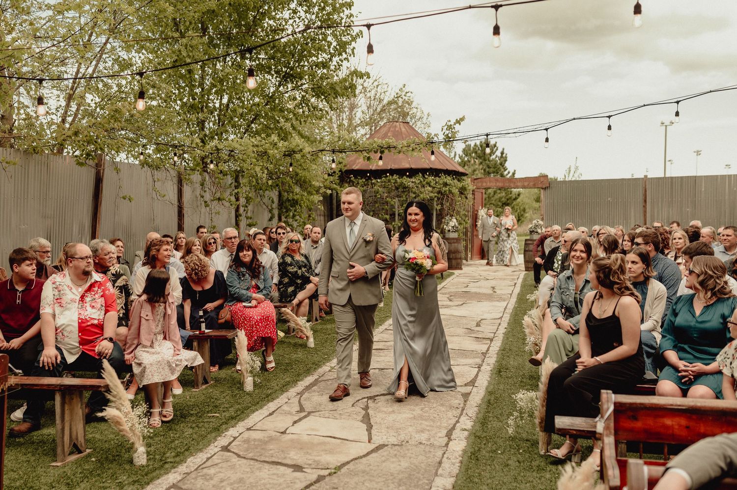 Outdoor wedding ceremony setup with string lights and guests seated in rows on a grassy hillside at sunset.