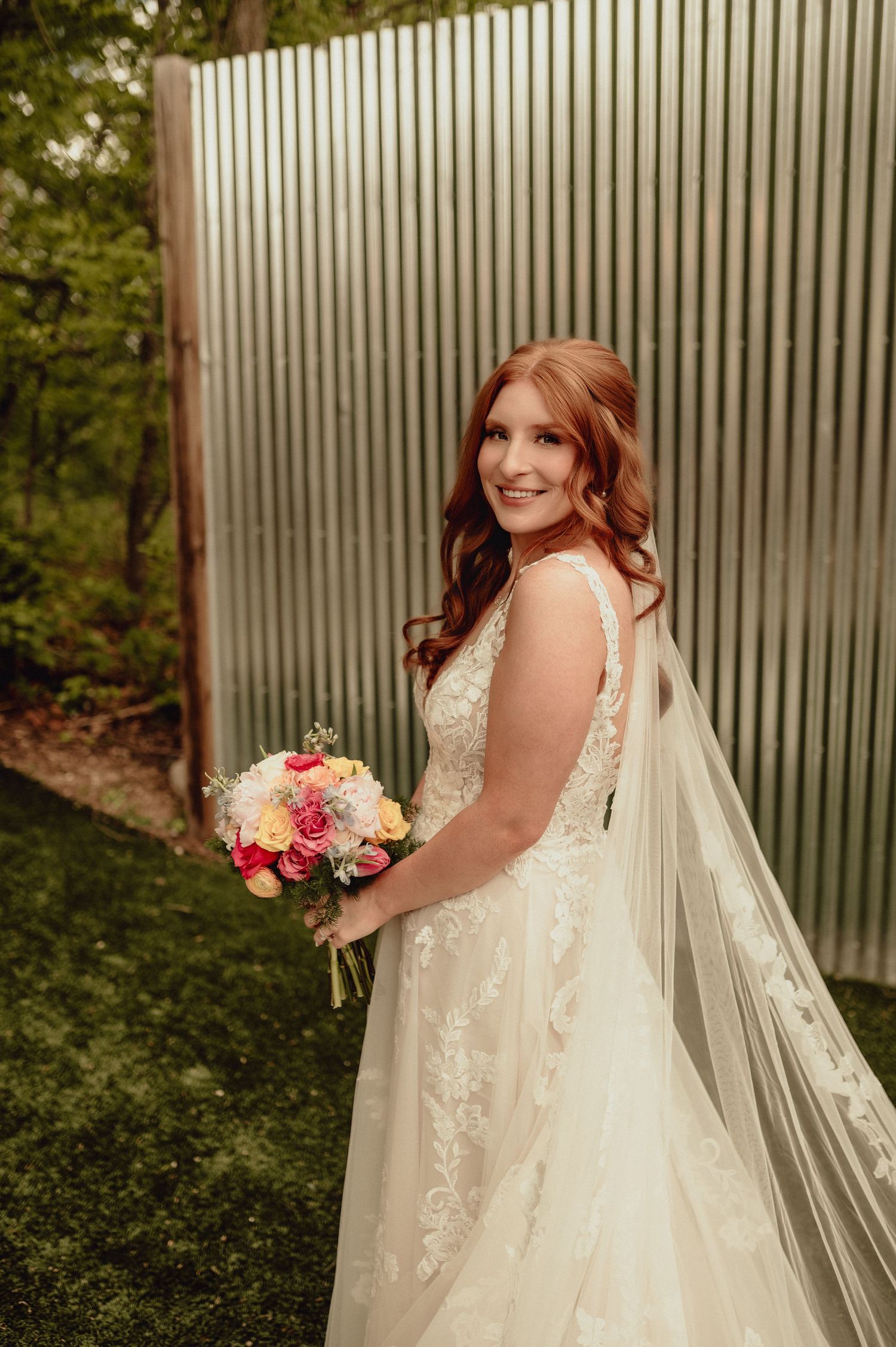 A bride in a lace wedding dress with long veil holds a pink and white bouquet against a corrugated metal backdrop.