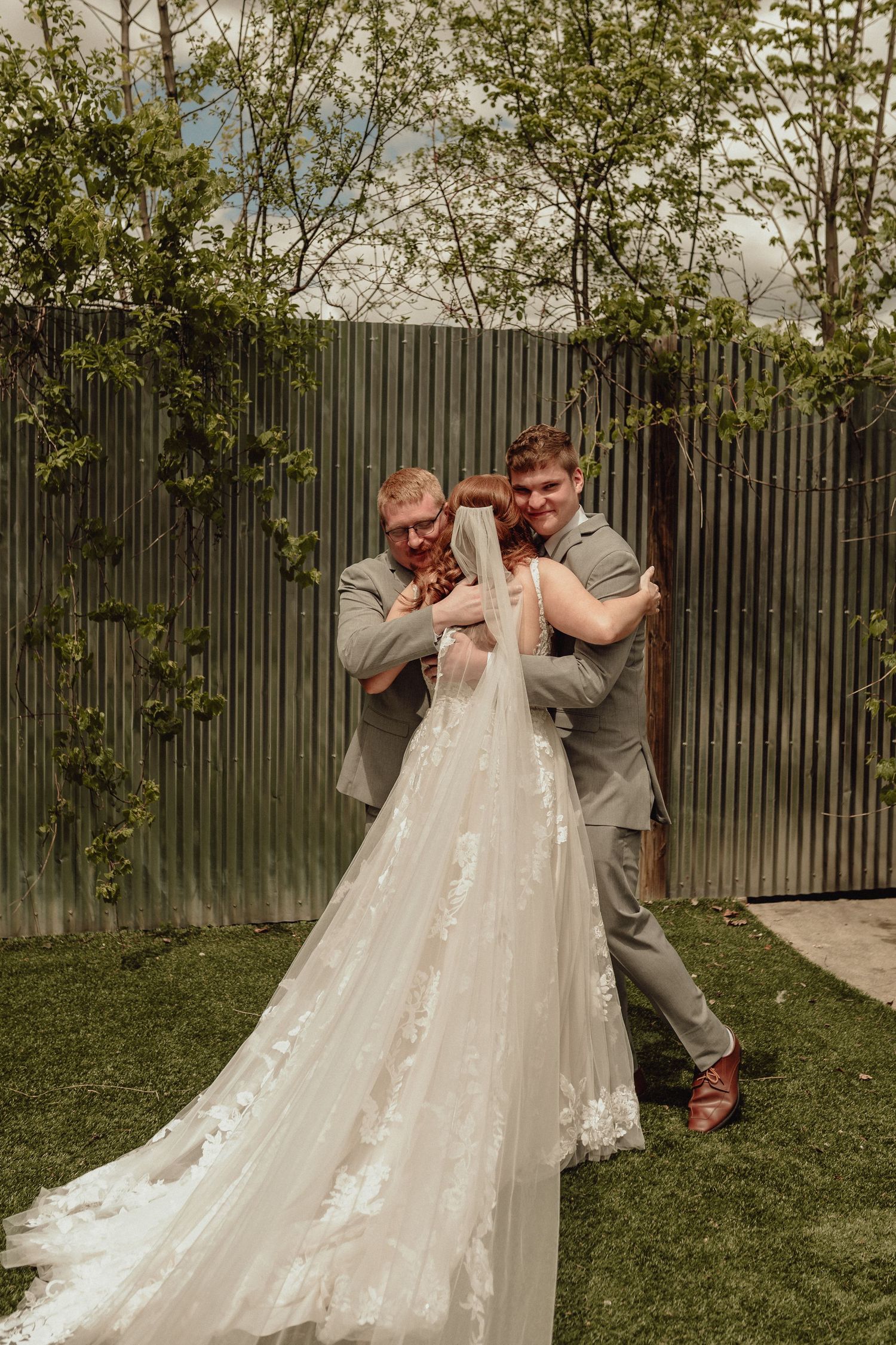 Two people in gray suits playfully pose with a flowing white wedding dress with lace details in an outdoor garden setting.
