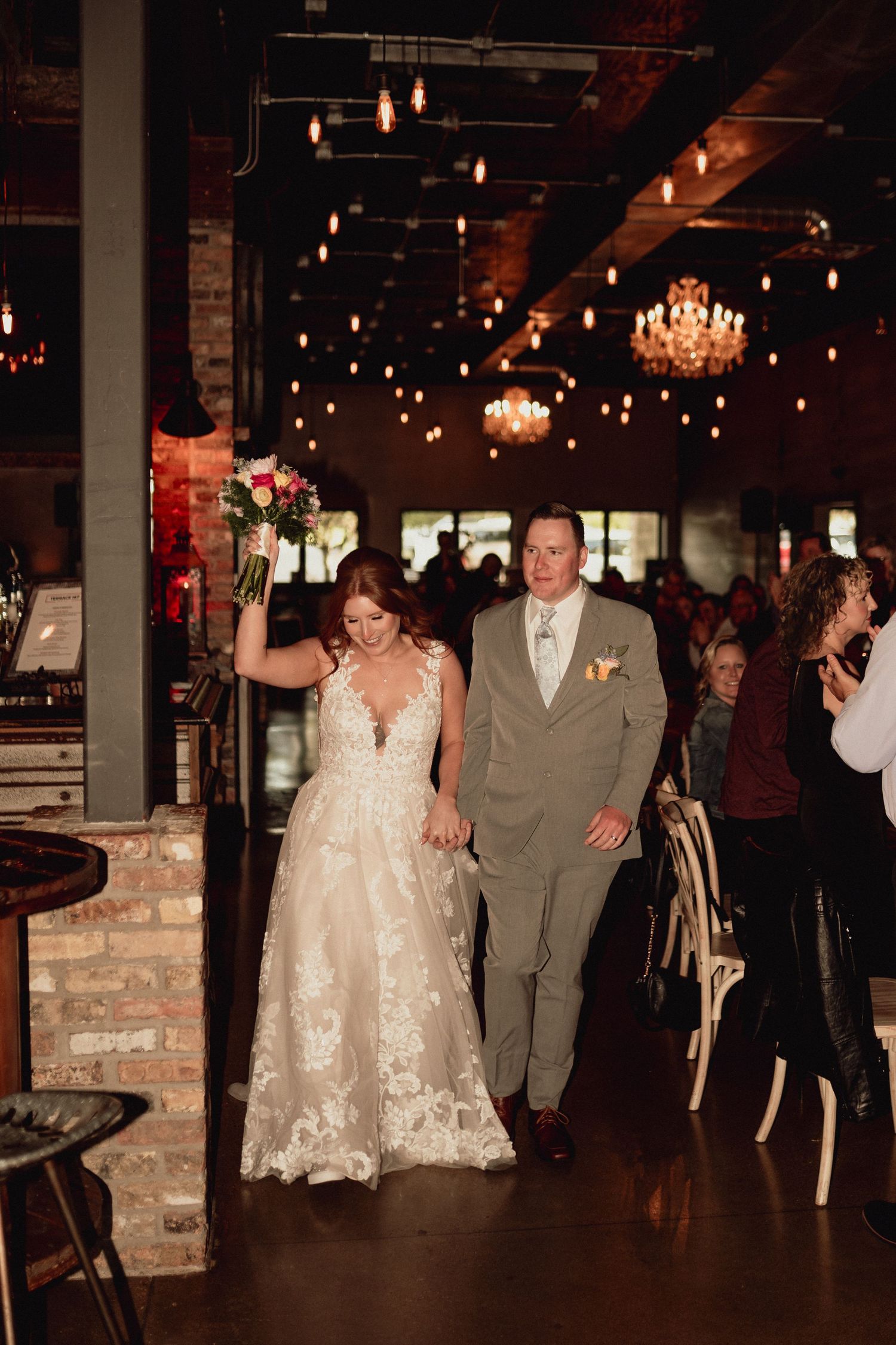 Wedding guests dance and celebrate at a dimly lit rustic venue with exposed brick walls and hanging lights.