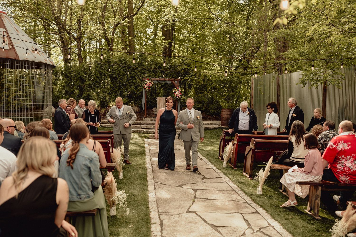 Wedding guests gather outdoors in a wooded setting for a casual celebration event.