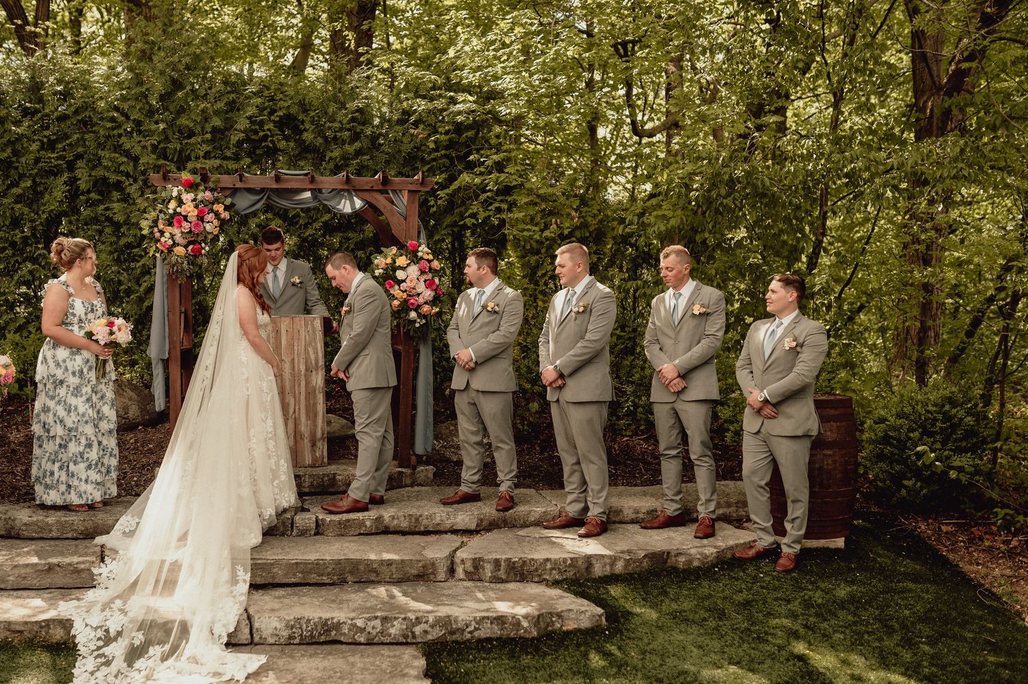 Wedding party stands together in a lush garden setting with stone steps and natural greenery backdrop.
