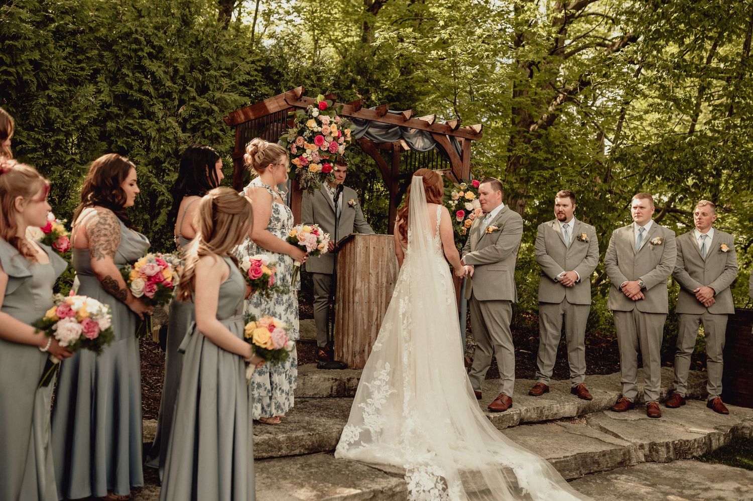 Outdoor wedding ceremony with bridesmaids in sage green dresses and groomsmen in grey suits under a rustic wooden arch.