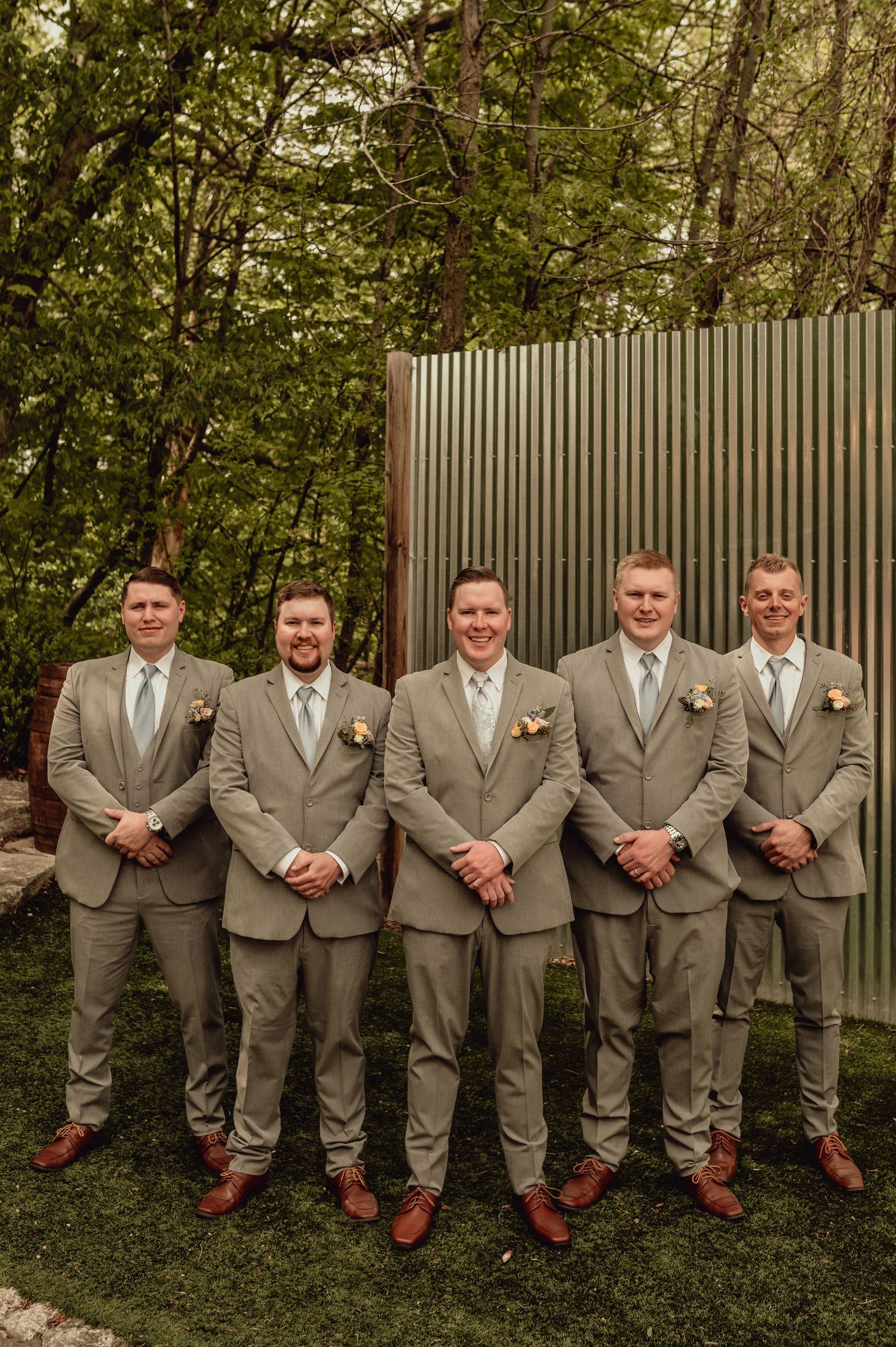 A group of groomsmen in matching tan suits and brown shoes pose casually against a wooden fence outdoors.