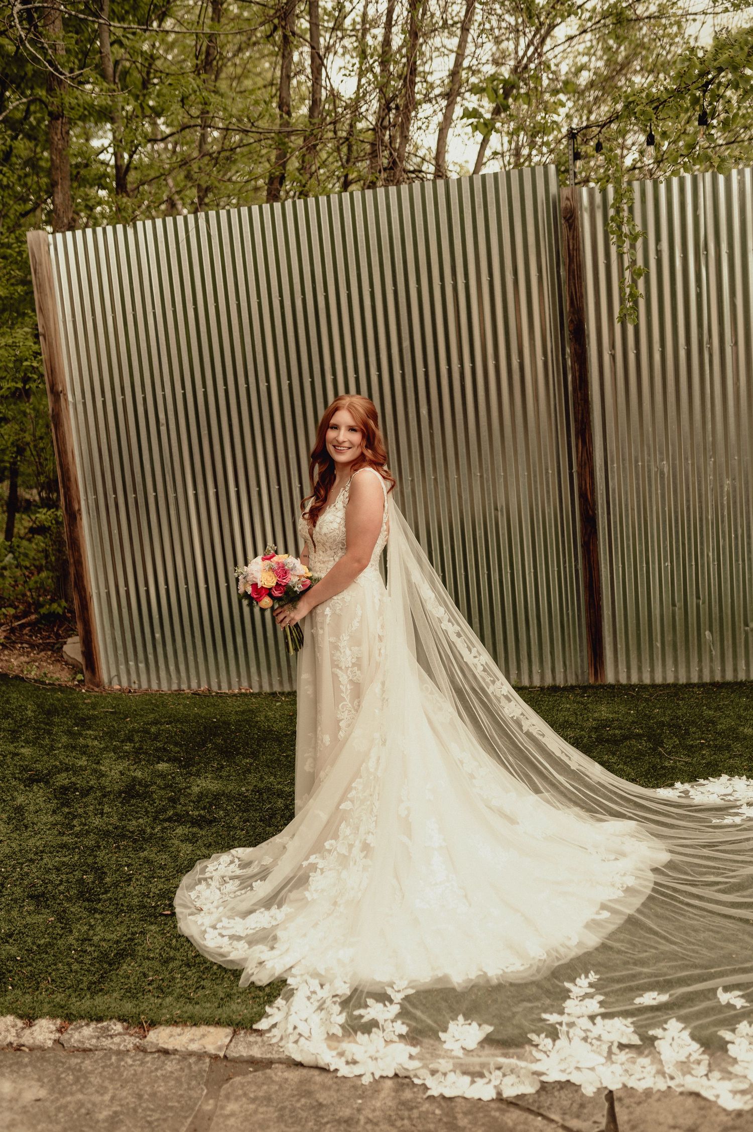 A bride in a stunning lace wedding dress with a long cathedral veil poses against a white fence in an outdoor garden setting.