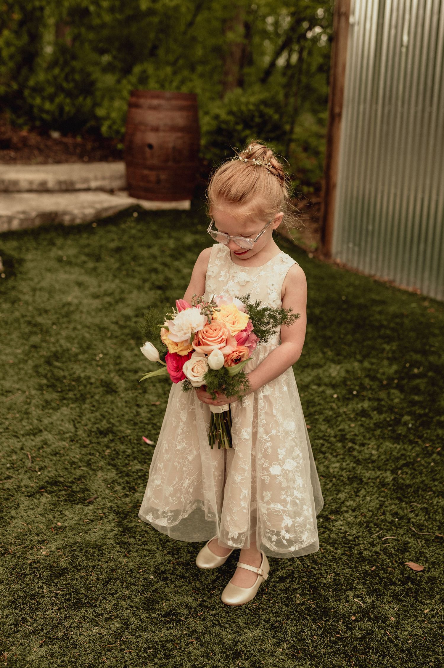 A flower girl in a white lace dress holds a colorful bouquet while sitting on green grass during a wedding.