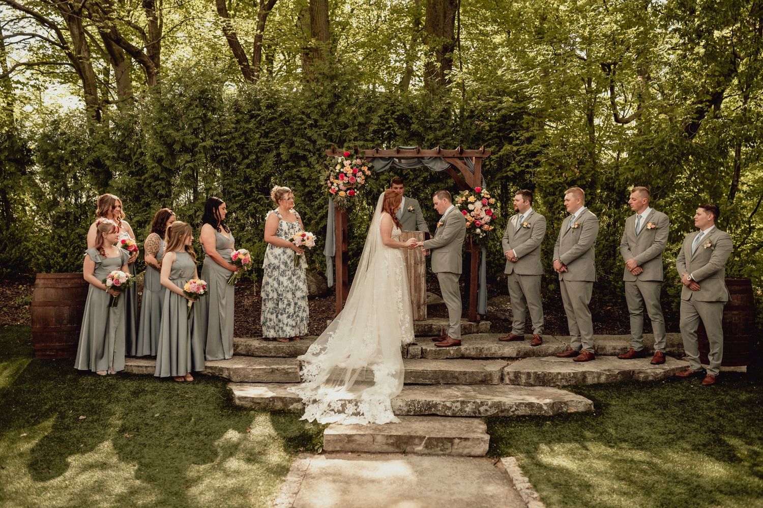 Wedding party members stand in a row outdoors under trees during a summer ceremony.