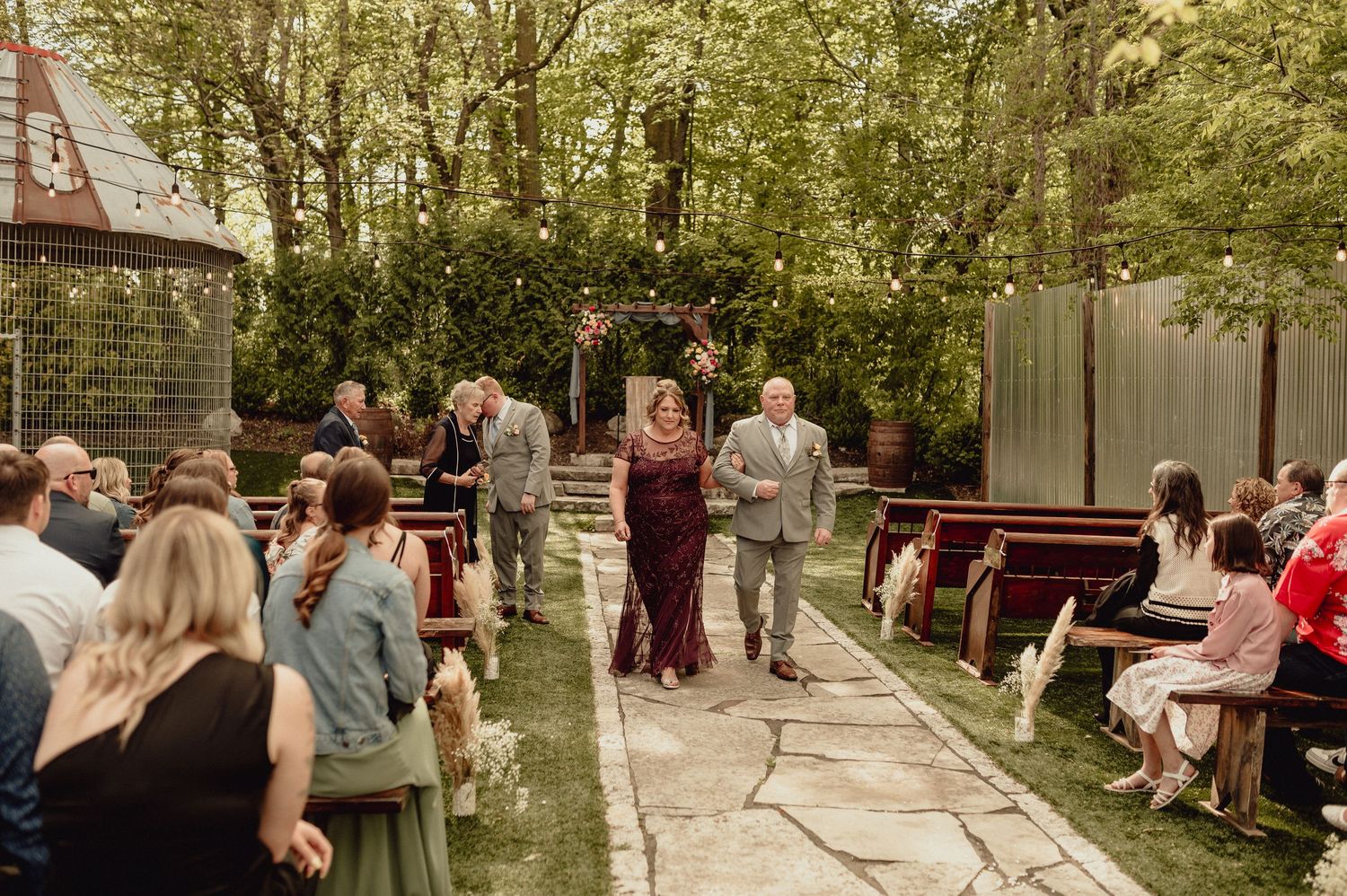 People gather outdoors at rustic wooden picnic tables surrounded by trees for a casual dining event.