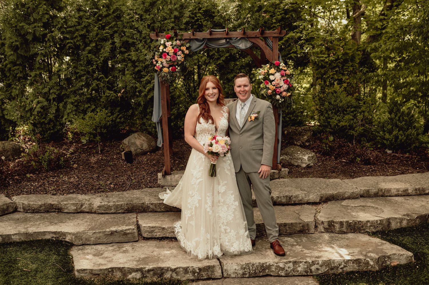 A couple poses together on a rustic stone wall surrounded by lush greenery and flowers during their wedding celebration.