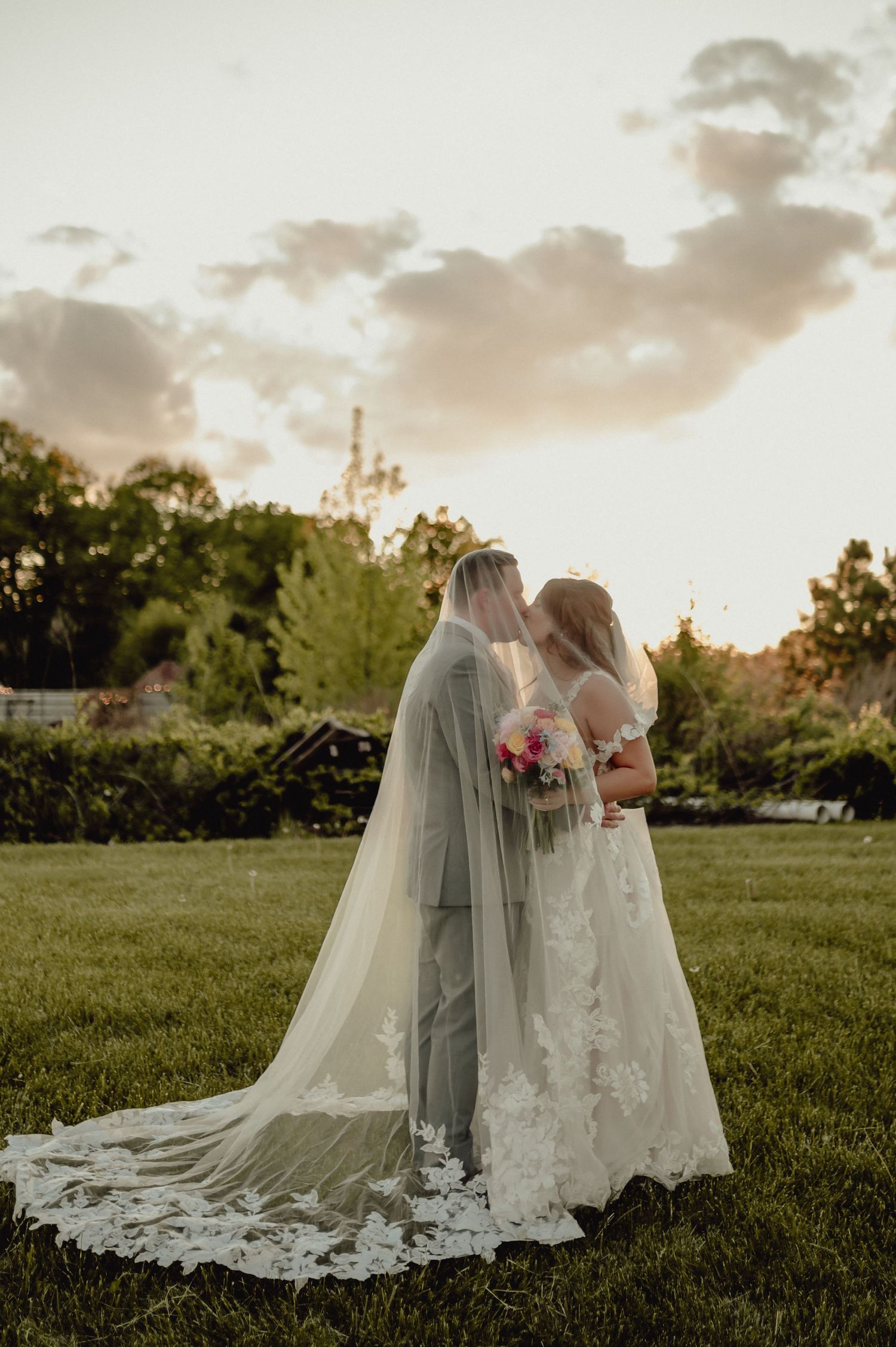 Two people in wedding attire embrace on a grassy field during golden hour sunset with flowing veils and lace dresses.