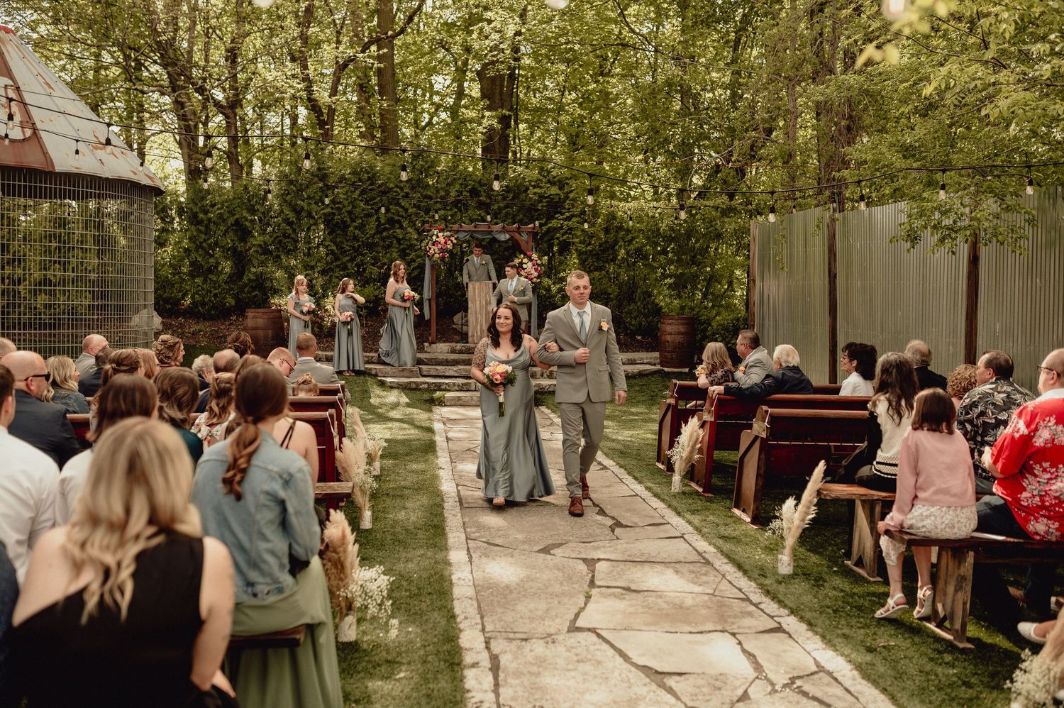 Wedding guests gather outdoors in a wooded setting for a rustic ceremony celebration.