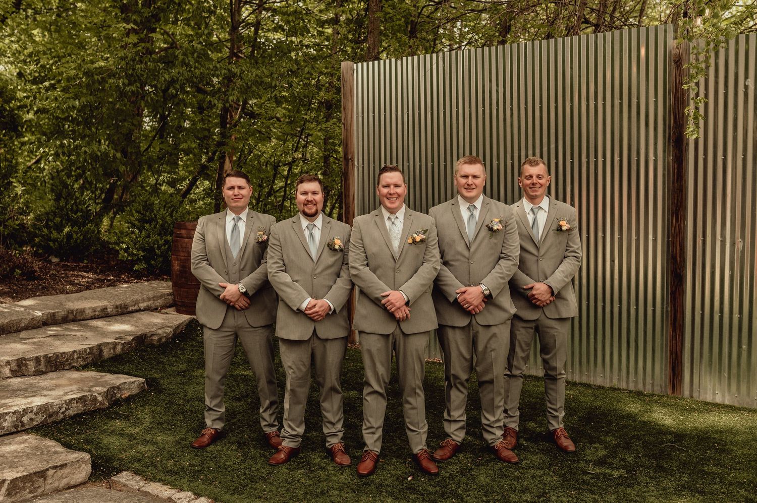 Groomsmen in matching gray suits pose together on outdoor stone steps during a wedding celebration.