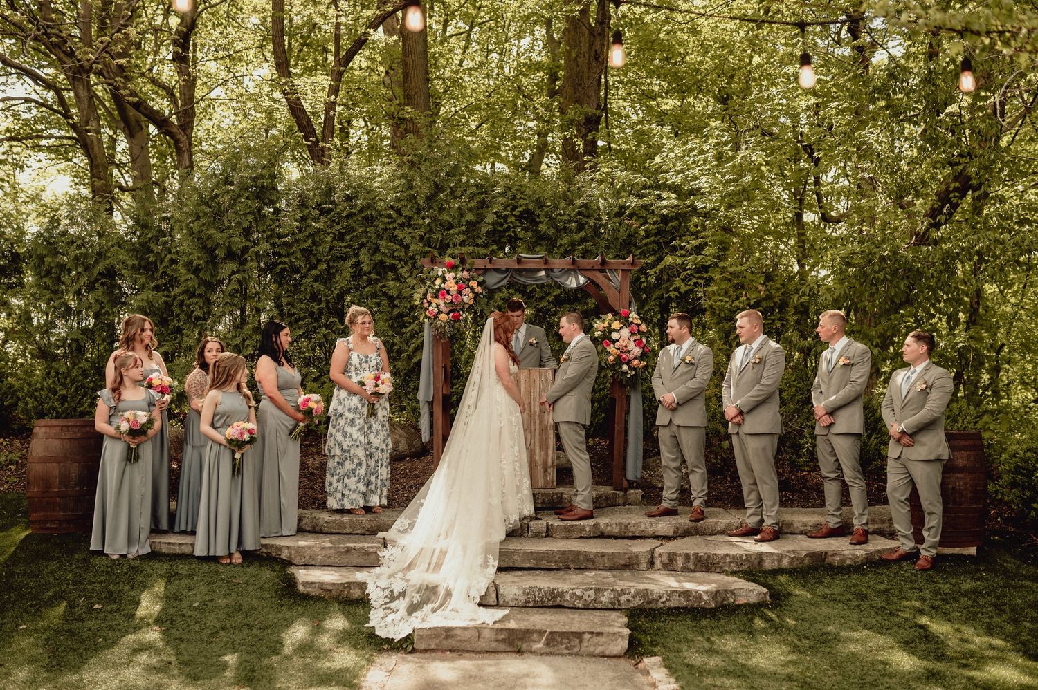 A wedding party stands in a row outdoors with bridesmaids in gray dresses and groomsmen in tan suits.