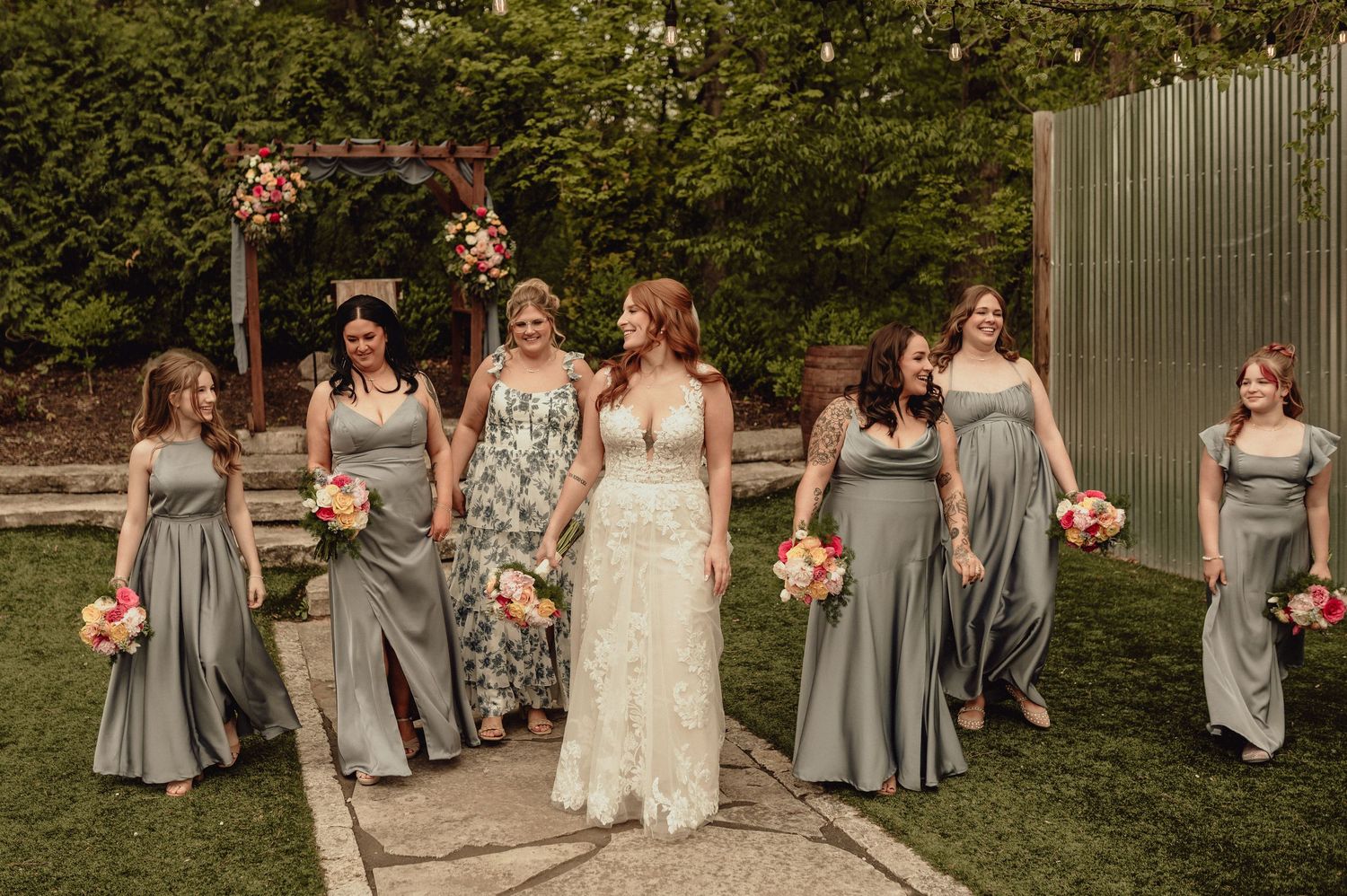Wedding party in gray bridesmaid dresses poses together outdoors with bouquets of pink flowers.