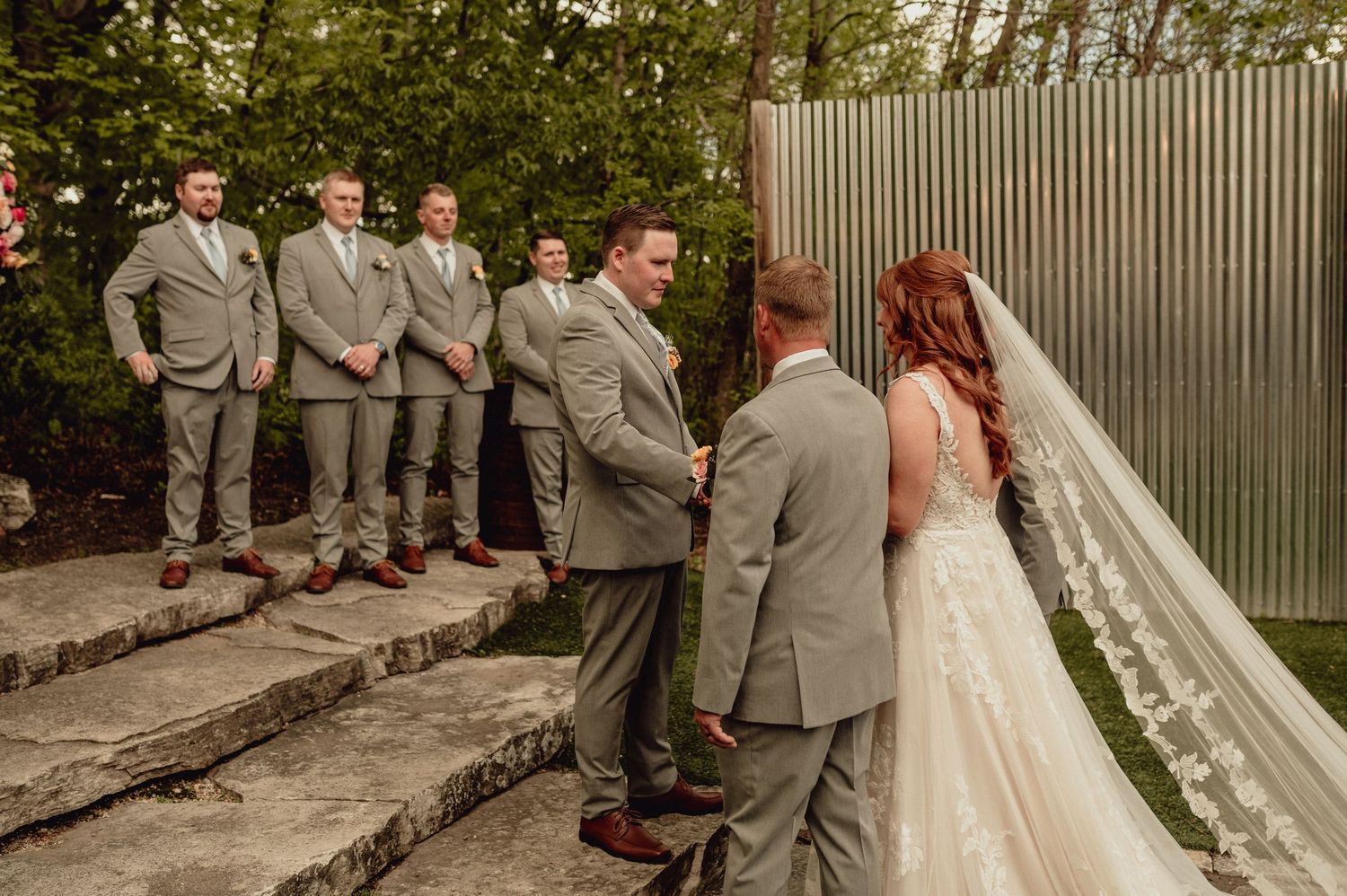 A wedding party in gray suits lines up along a garden wall during an outdoor ceremony.