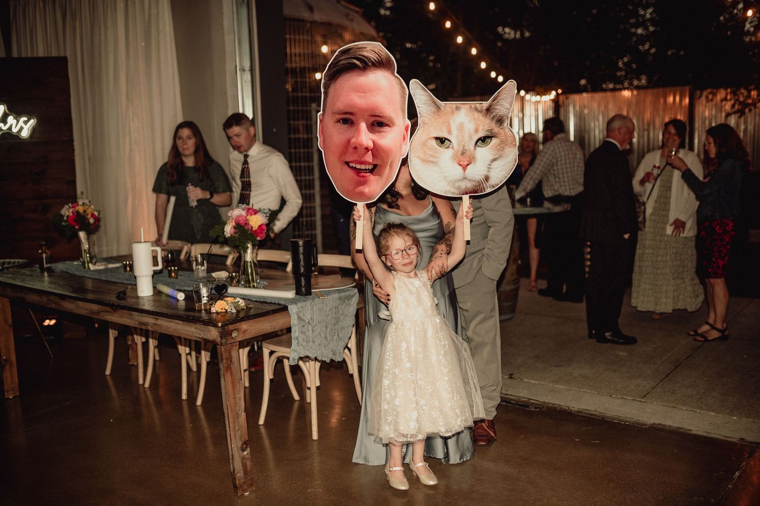 A person and a child in a light dress dance together at a wedding reception with string lights overhead.