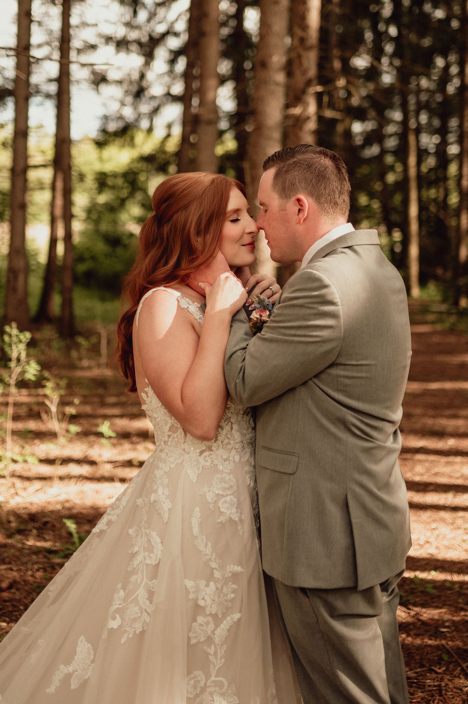 A romantic wedding couple embraces in a sunlit forest setting with pine trees and fallen leaves.