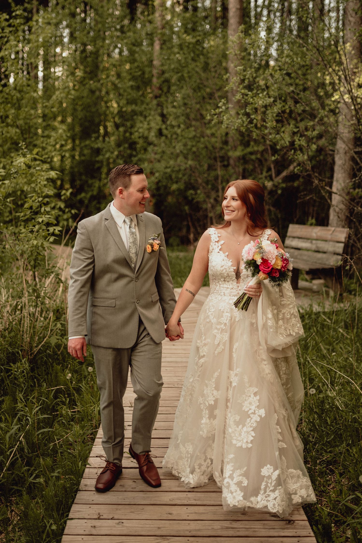 A couple in wedding attire holds hands while walking along a wooden boardwalk path surrounded by lush greenery.