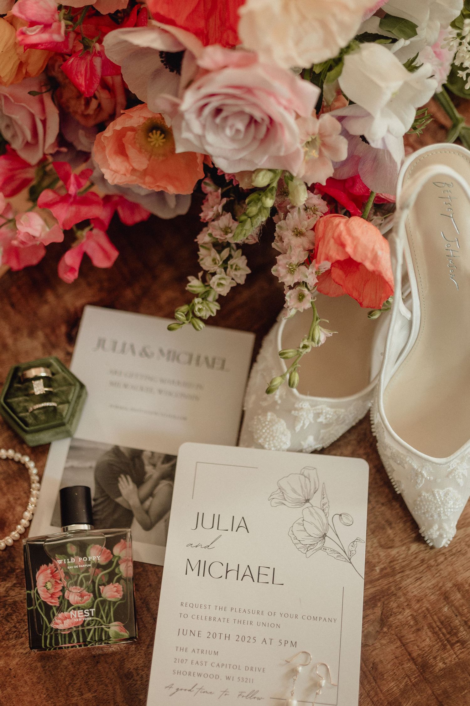 Wedding stationery and white bridal shoes arranged with pink and white flowers on rustic wooden table.