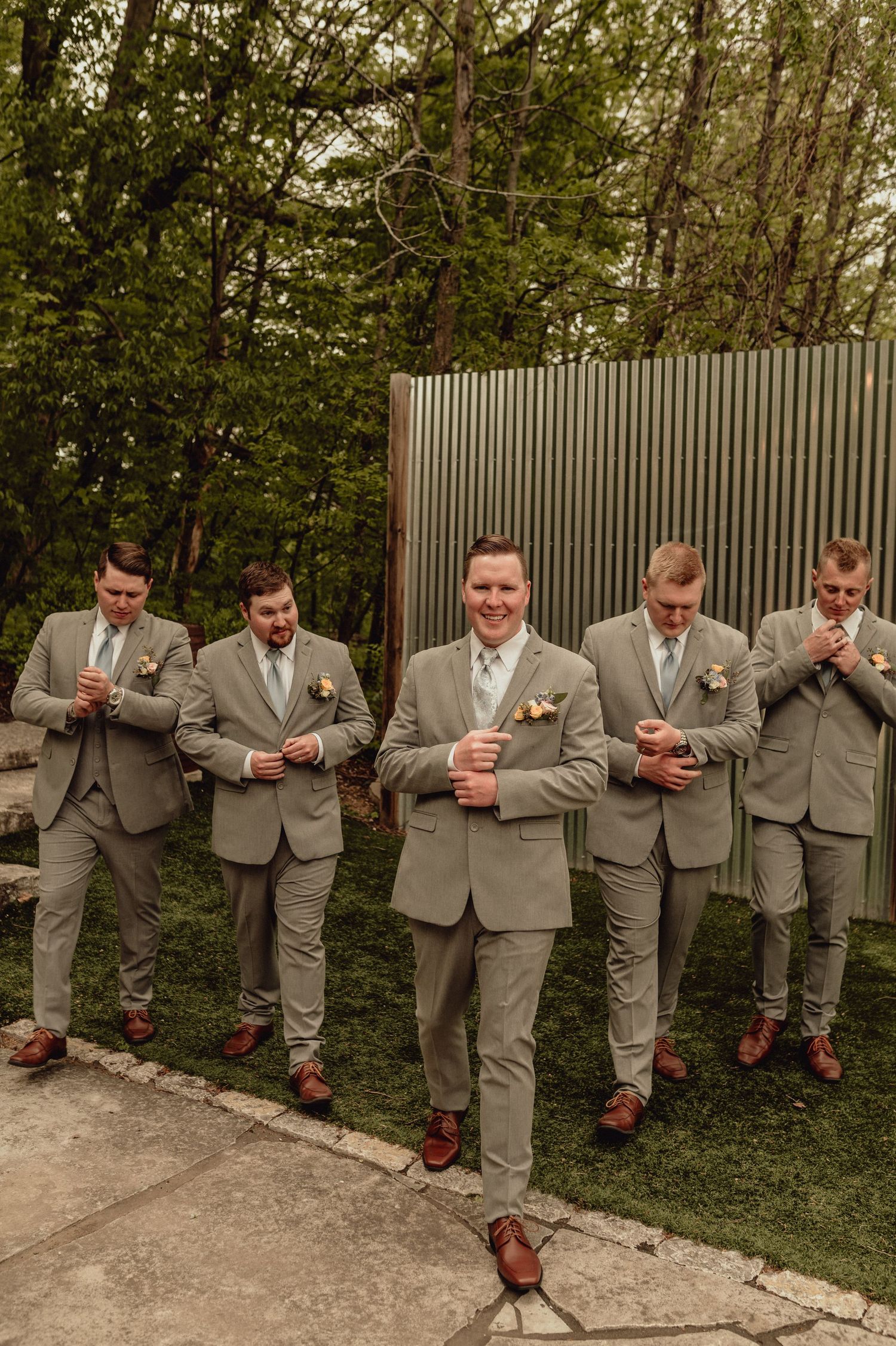 A group in matching gray suits and brown shoes perform a sequence of playful poses against a garden wall.