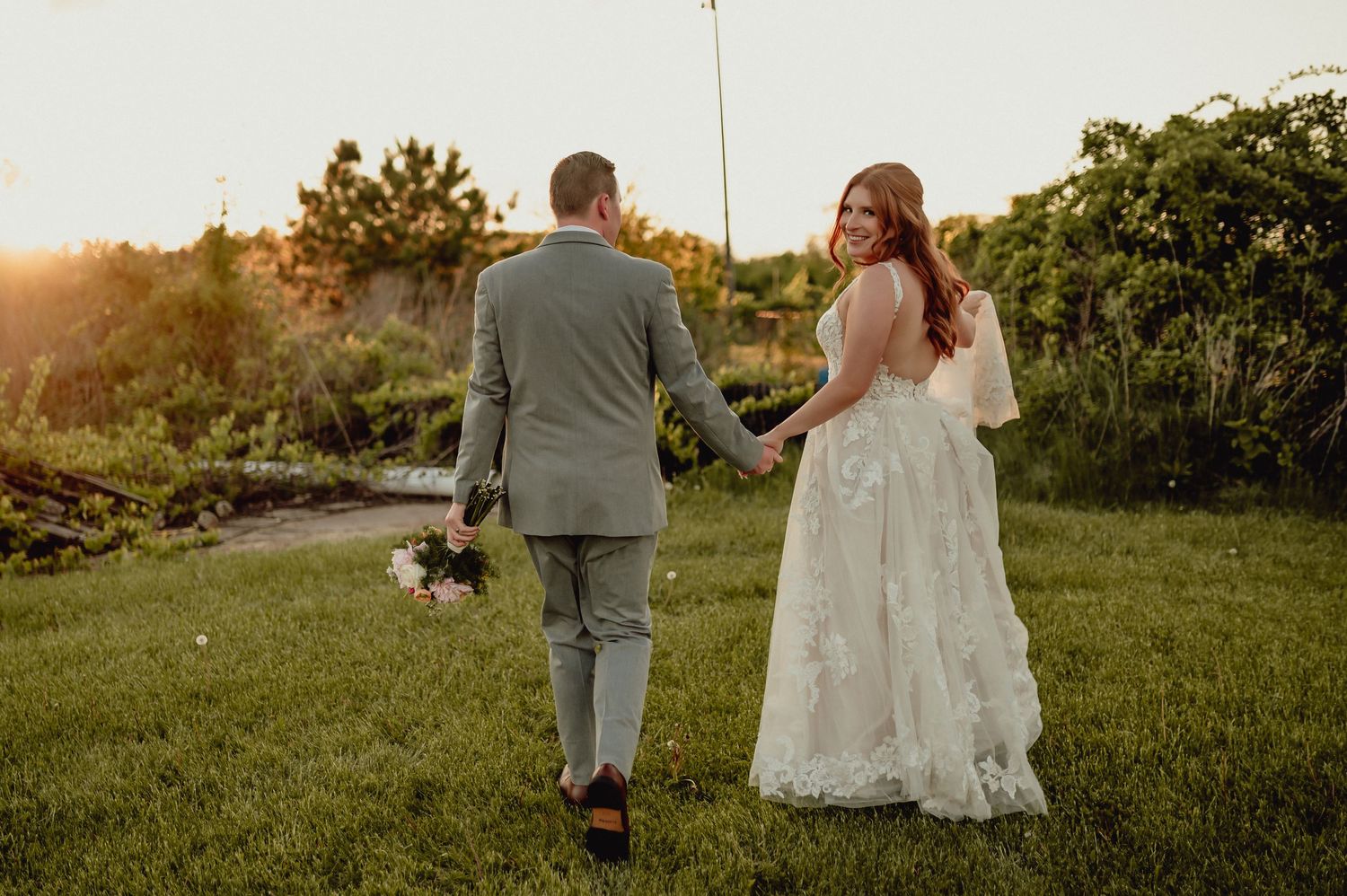 A couple walks hand in hand through a sunlit meadow at sunset in their wedding attire.