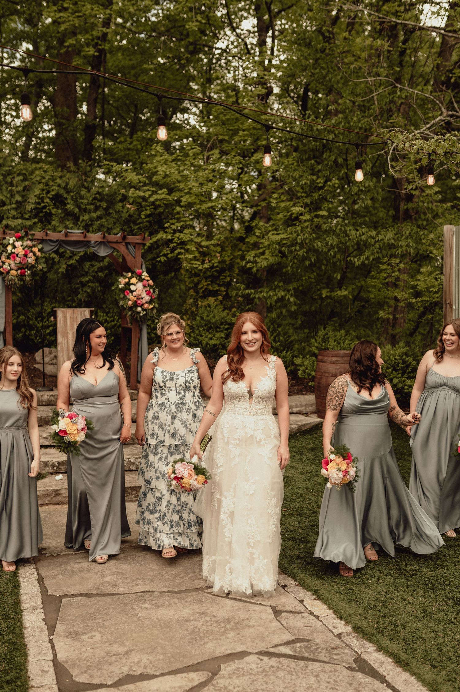 A wedding party poses outdoors with string lights and hanging floral decorations, bridesmaids wearing gray and floral dresses.