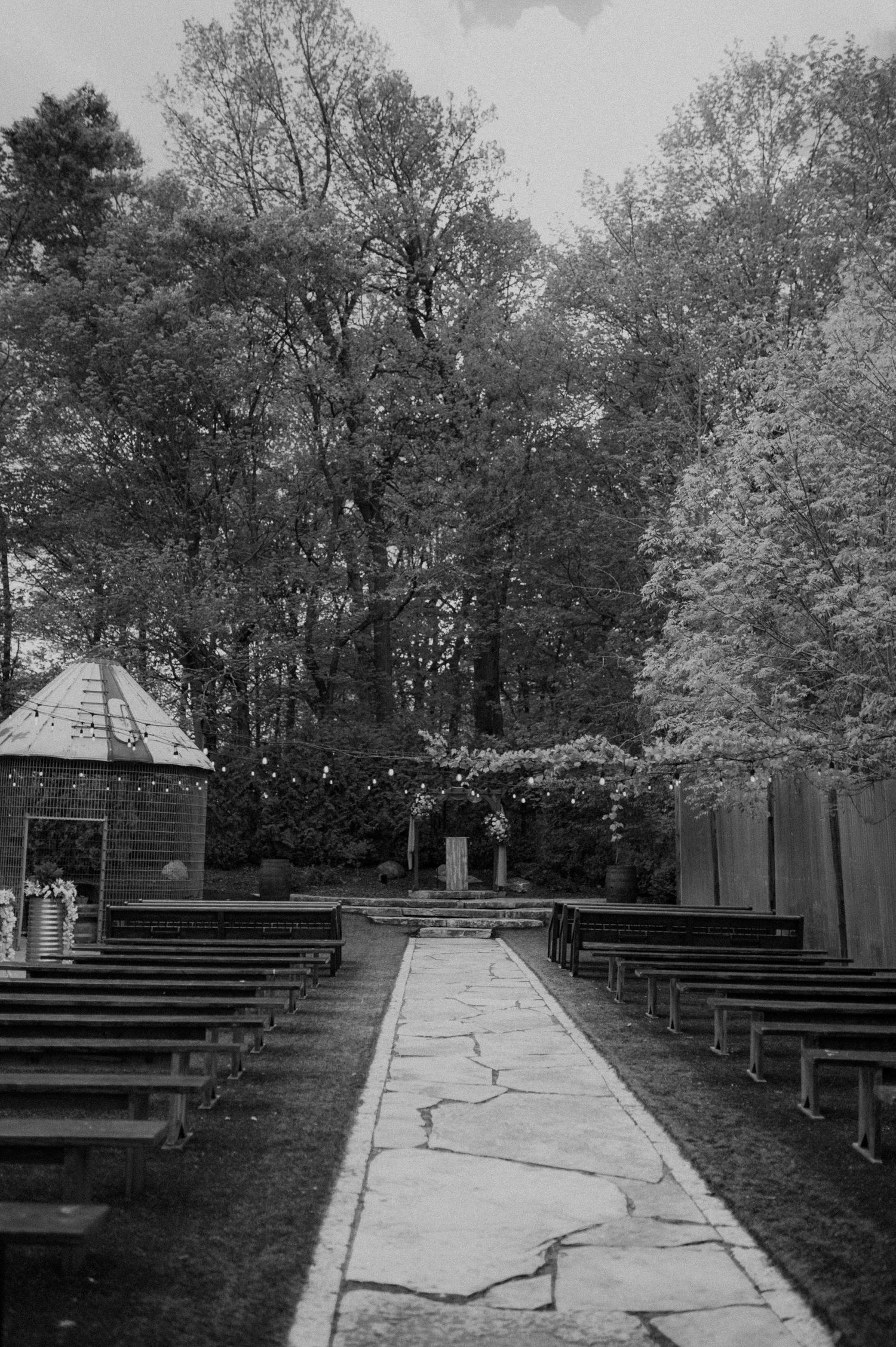 A black and white photograph of an outdoor chapel with wooden benches and a stone pathway leading to a small gazebo.