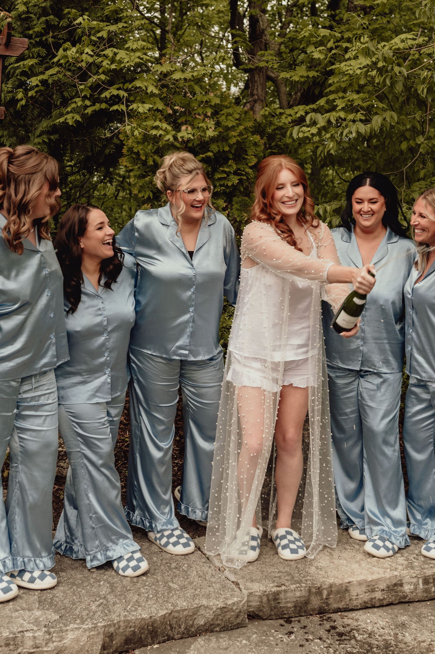 Wedding celebration with attendants in matching silk pajamas sharing champagne outdoors.