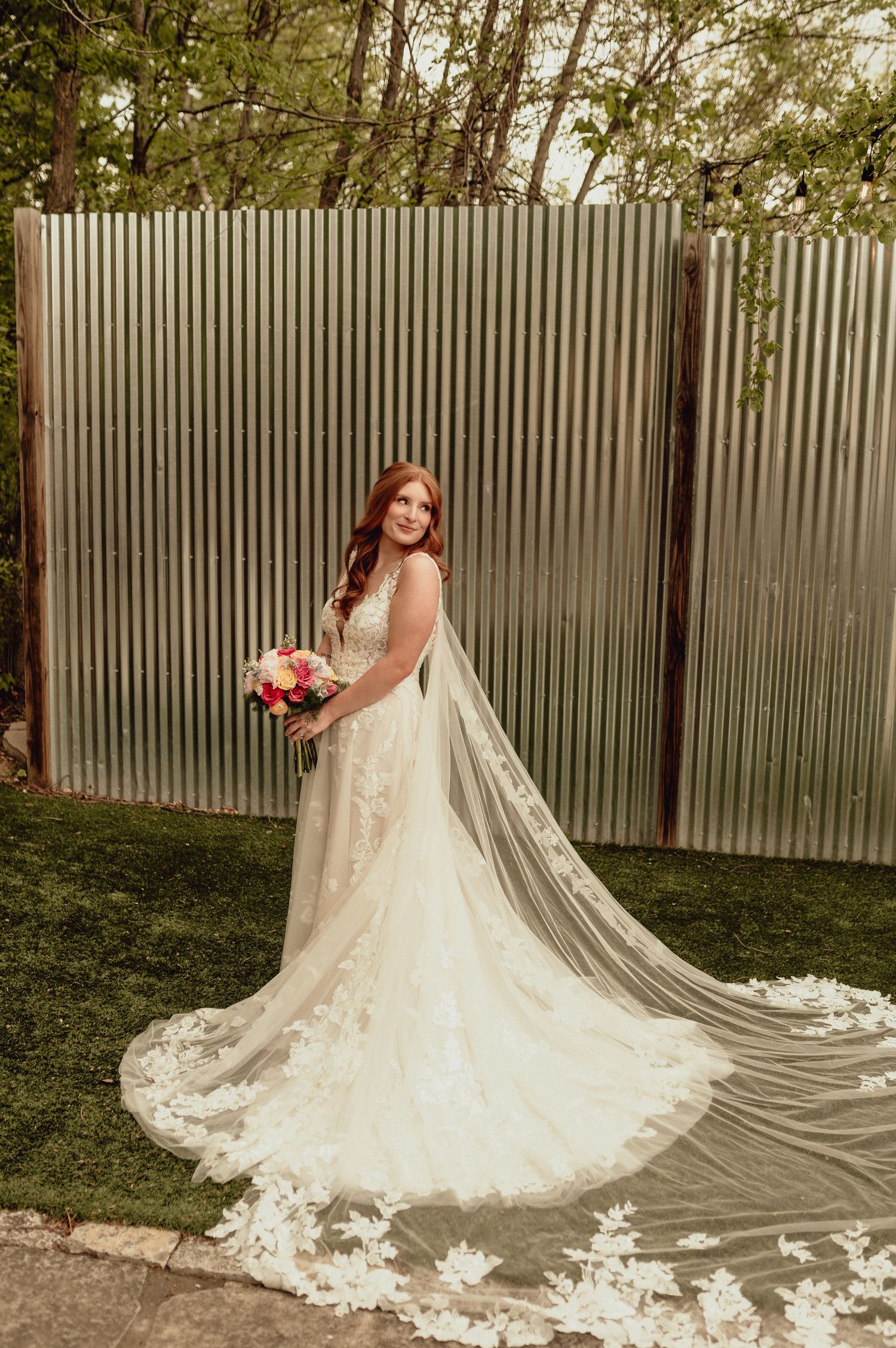 A bride in a flowing lace wedding gown with cathedral veil poses in a garden setting against a white fence.