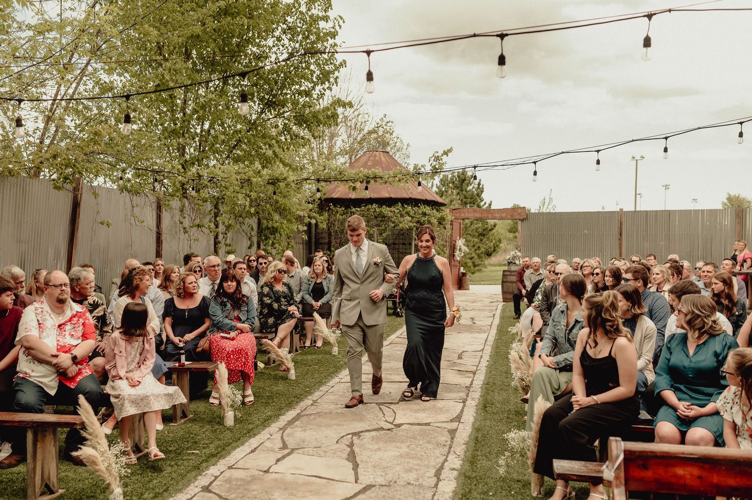 String lights illuminate an outdoor wedding reception with guests mingling and dancing on a lawn.