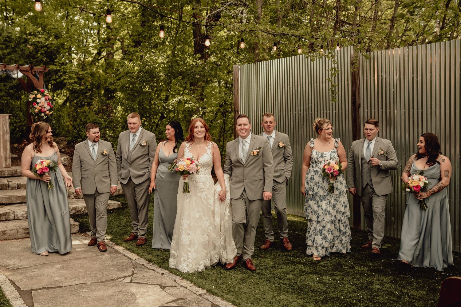 Wedding party in gray and floral dresses poses together in an outdoor garden setting with natural greenery.