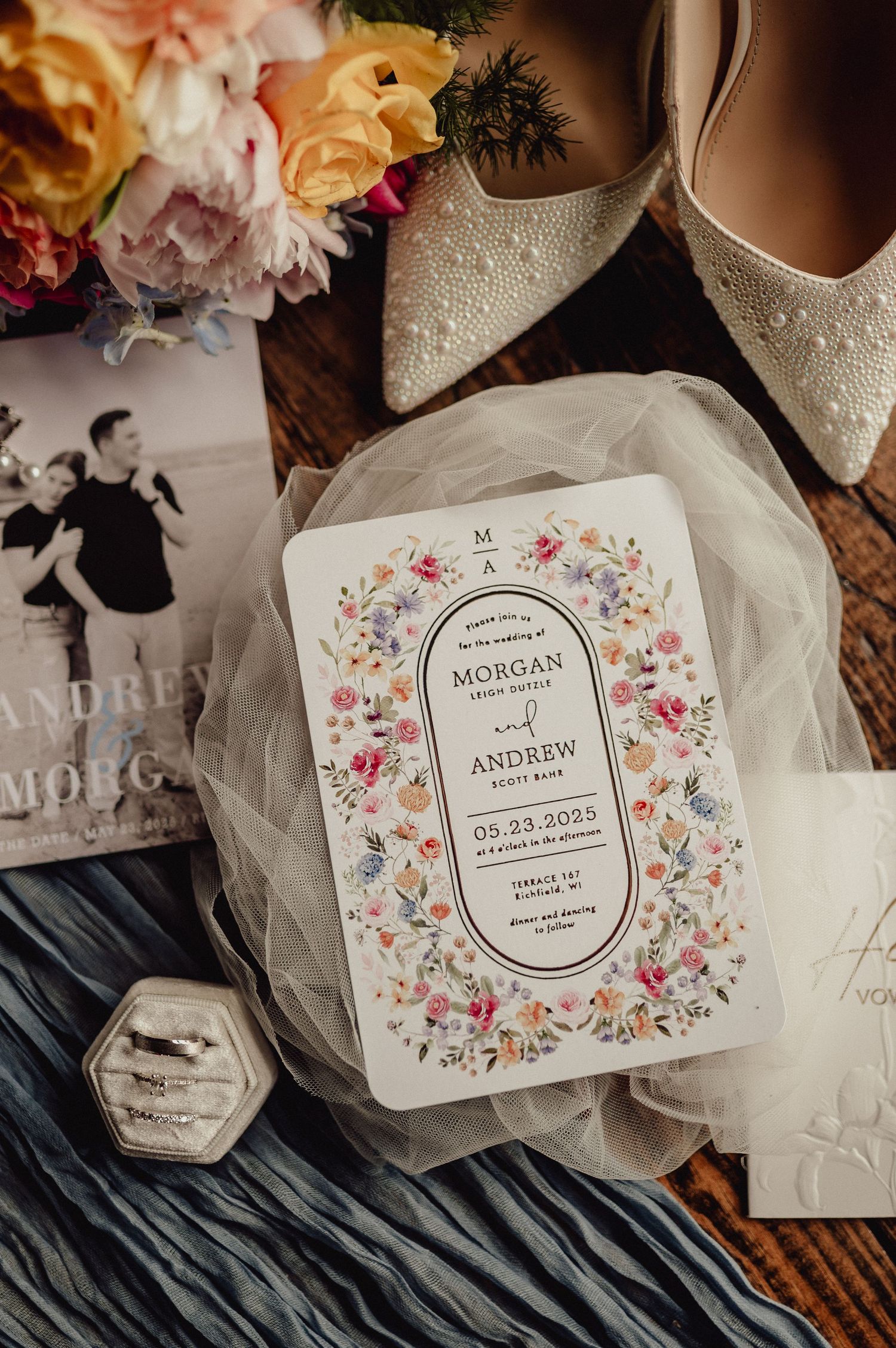 Wedding details including floral invitations, white shoes, and delicate veil arranged on dark wooden surface.