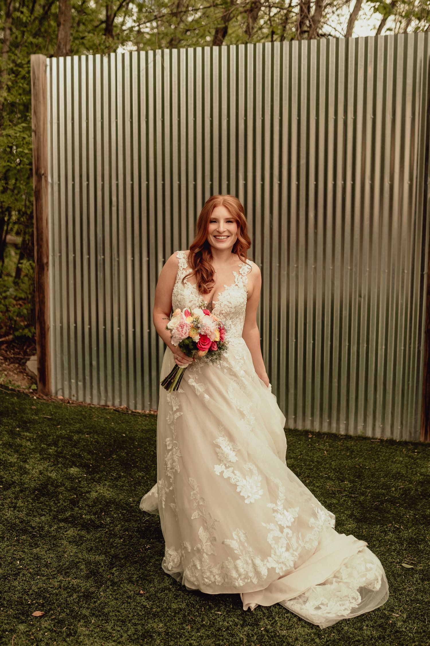 Bride in lace wedding dress poses against corrugated metal wall in outdoor garden setting with pink floral bouquet.