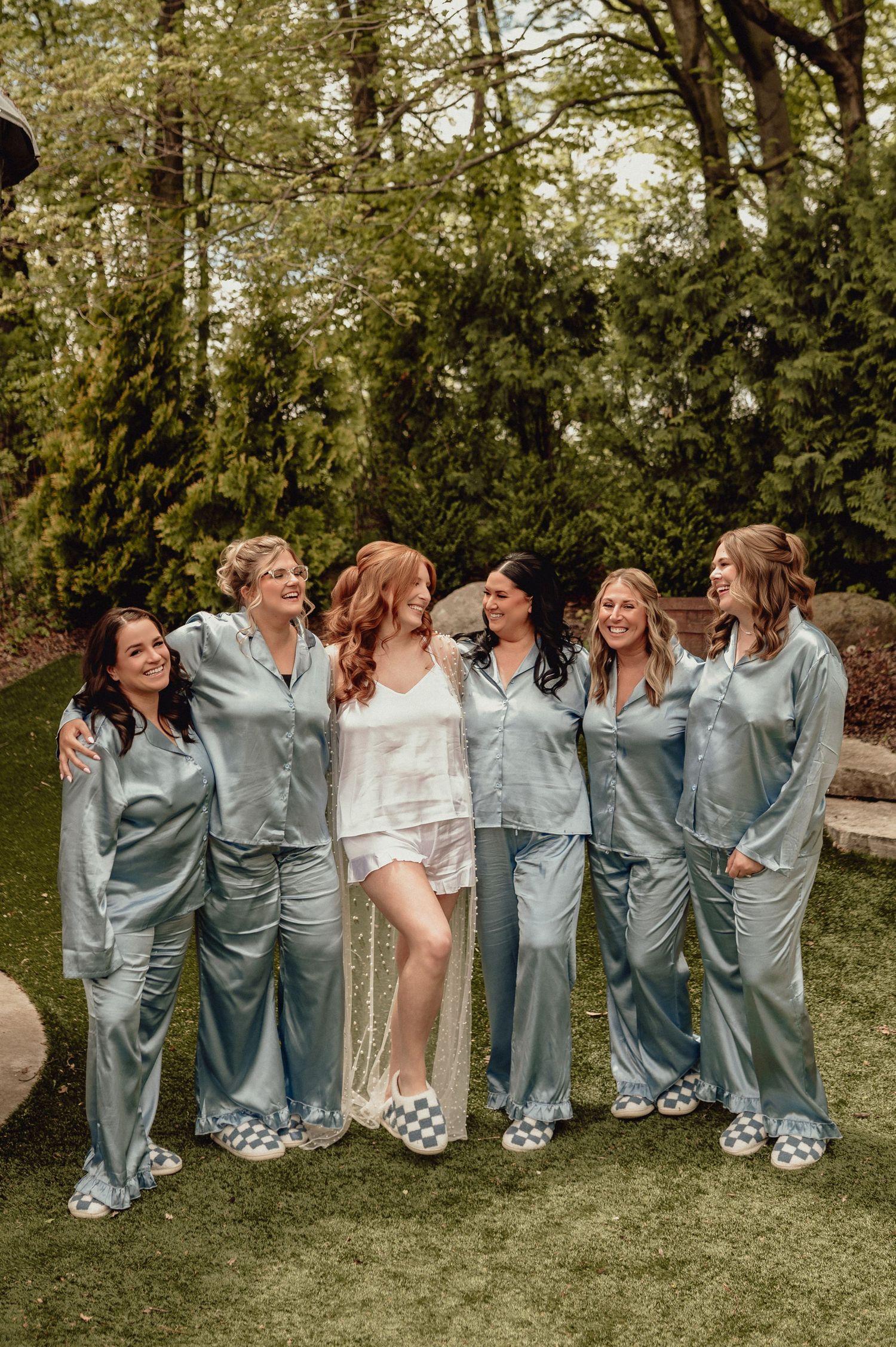 Group wearing matching blue satin pajamas poses together outdoors in a garden setting for a celebratory photoshoot.