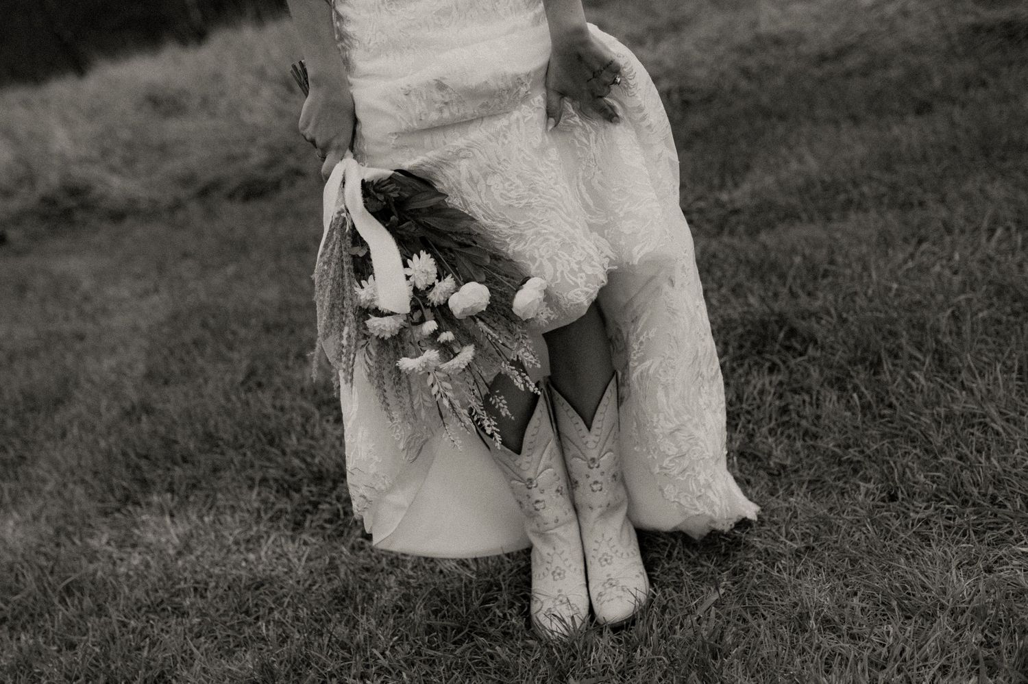 Black and white photograph of a bride in a white dress holding a bouquet while sitting on grass.