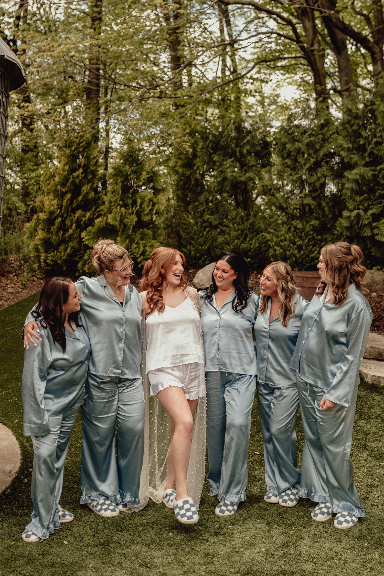 A bridal party in matching gray satin robes and fuzzy slippers poses playfully together on a grassy lawn.