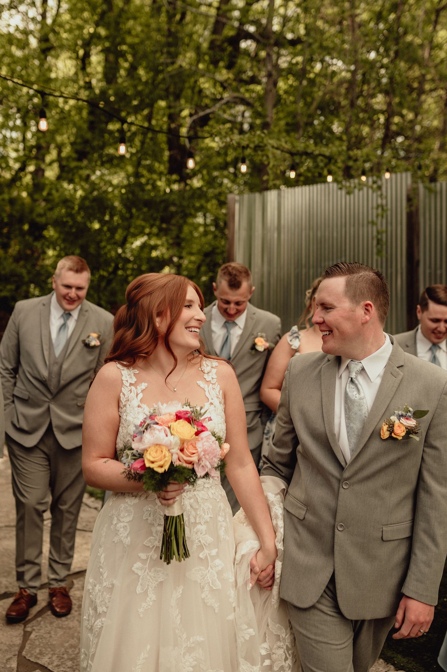 Wedding party in gray suits and ivory dress share candid laughing moments during outdoor photo session.