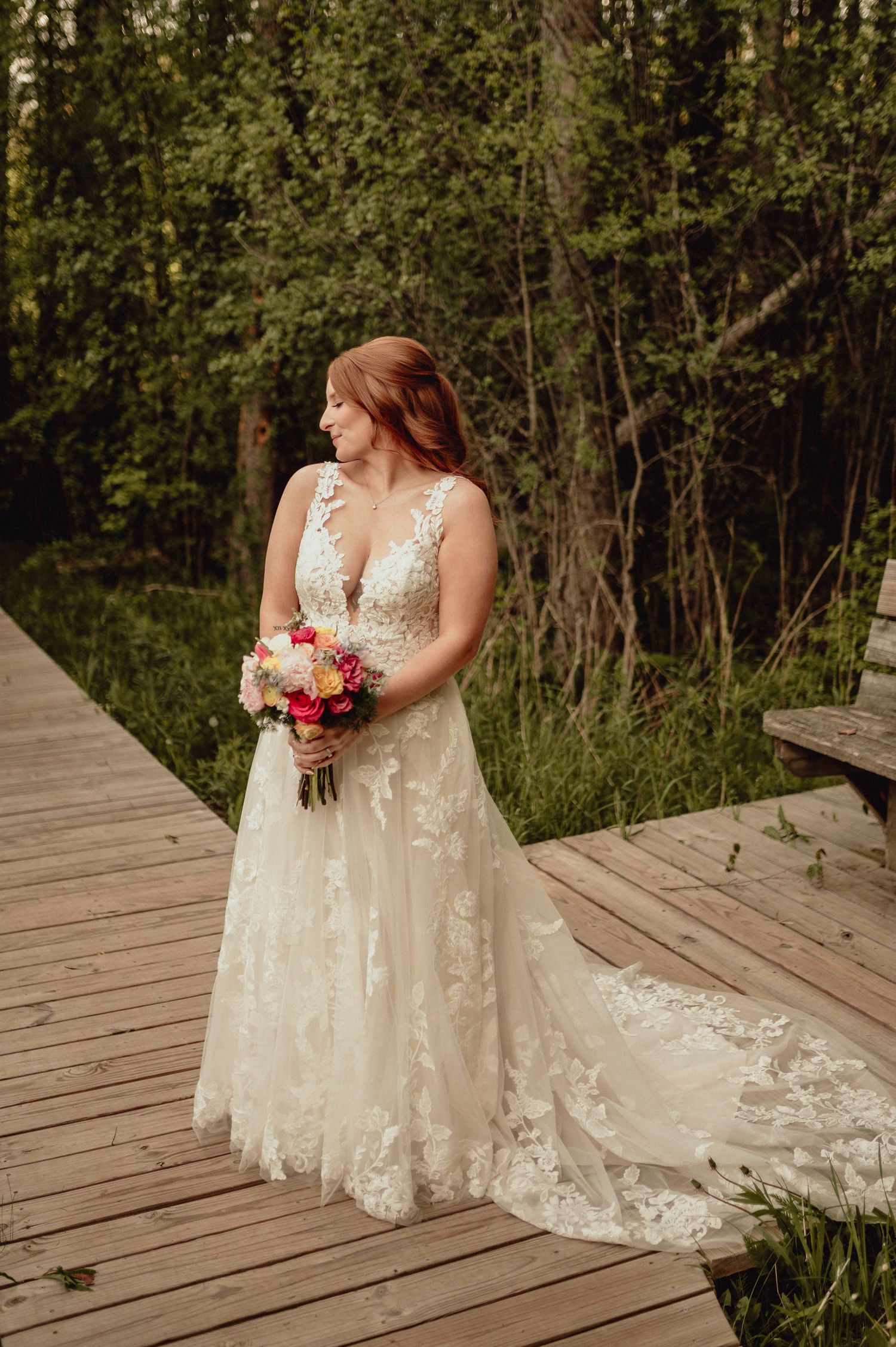 A bride in a lace wedding dress with floral appliques poses on a wooden deck surrounded by greenery.
