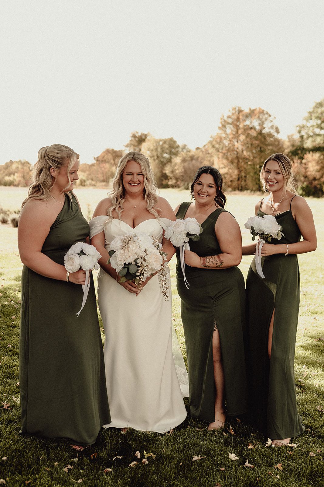 Wedding party in olive green dresses and white gown posing together in a sunlit field.