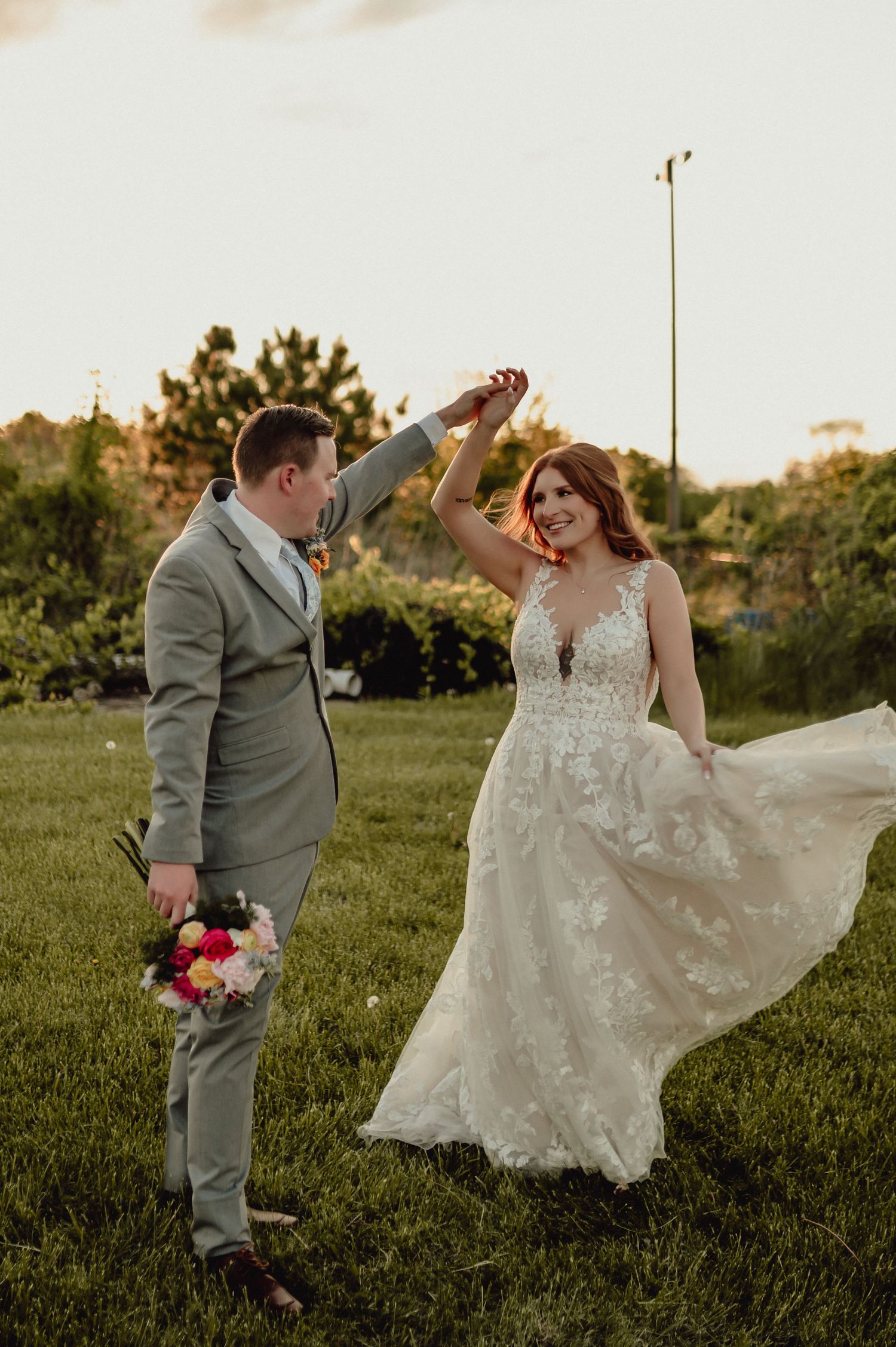 A couple dances playfully in a grassy field at sunset, with the bride's lace dress flowing elegantly in the breeze.