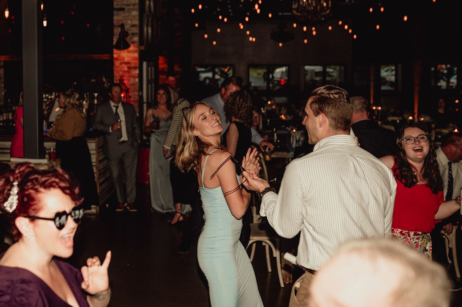 People dancing and socializing at a dimly lit party or reception with festive lighting in the background.