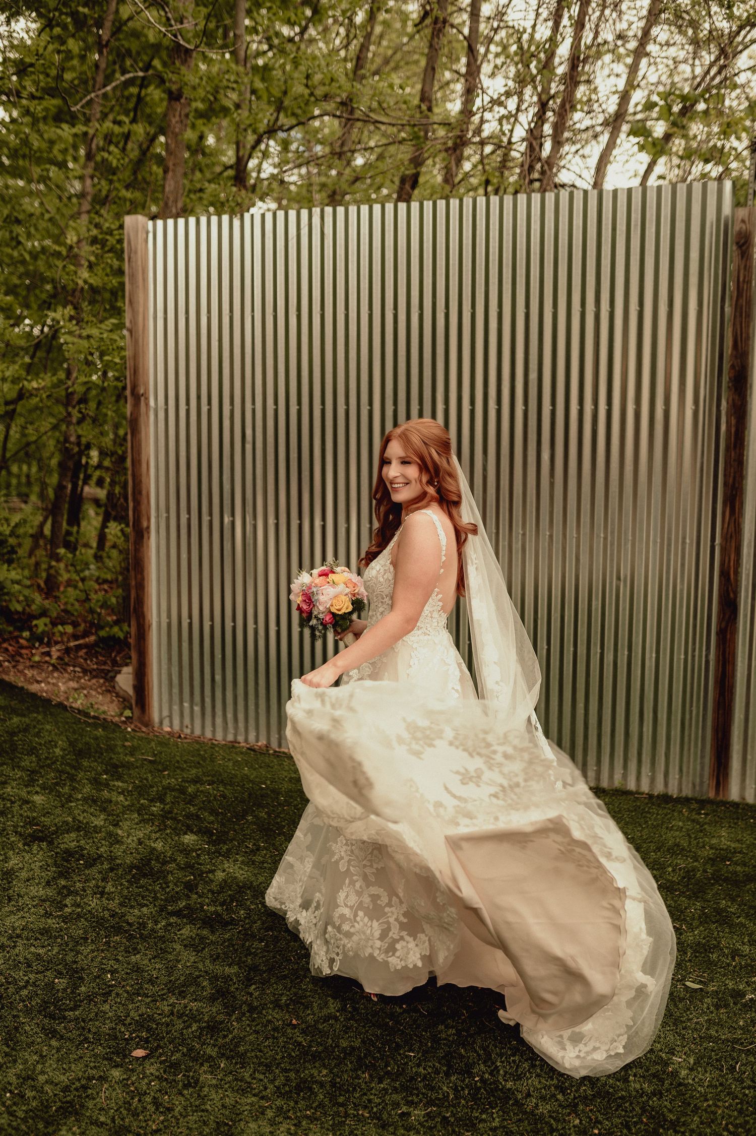 A bride in a flowing lace wedding dress twirls gracefully in front of a metal fence surrounded by green foliage.