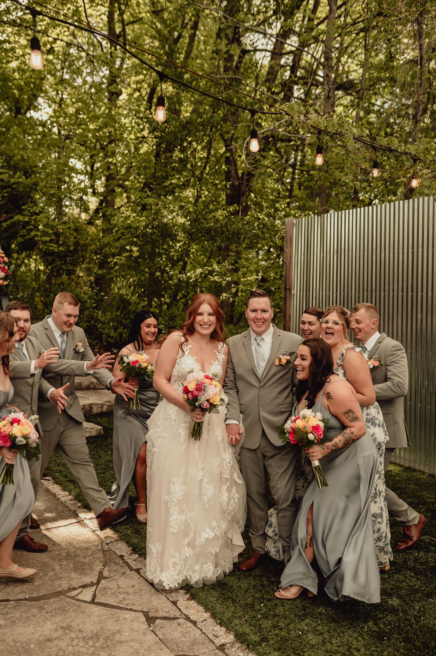 Wedding party shares candid laughs and poses against green foliage and a wooden fence.