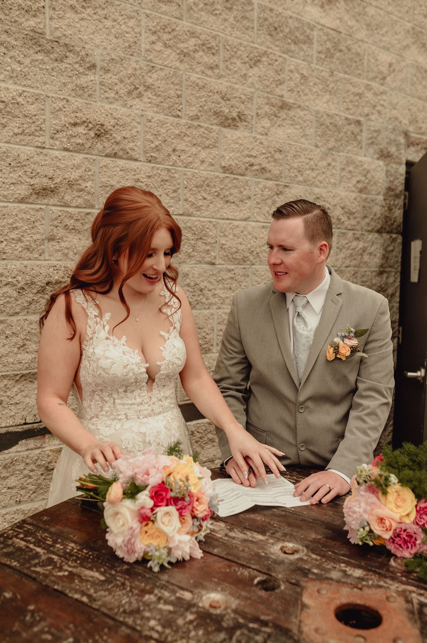 A couple in wedding attire share an intimate moment while sitting at a rustic wooden table outdoors.