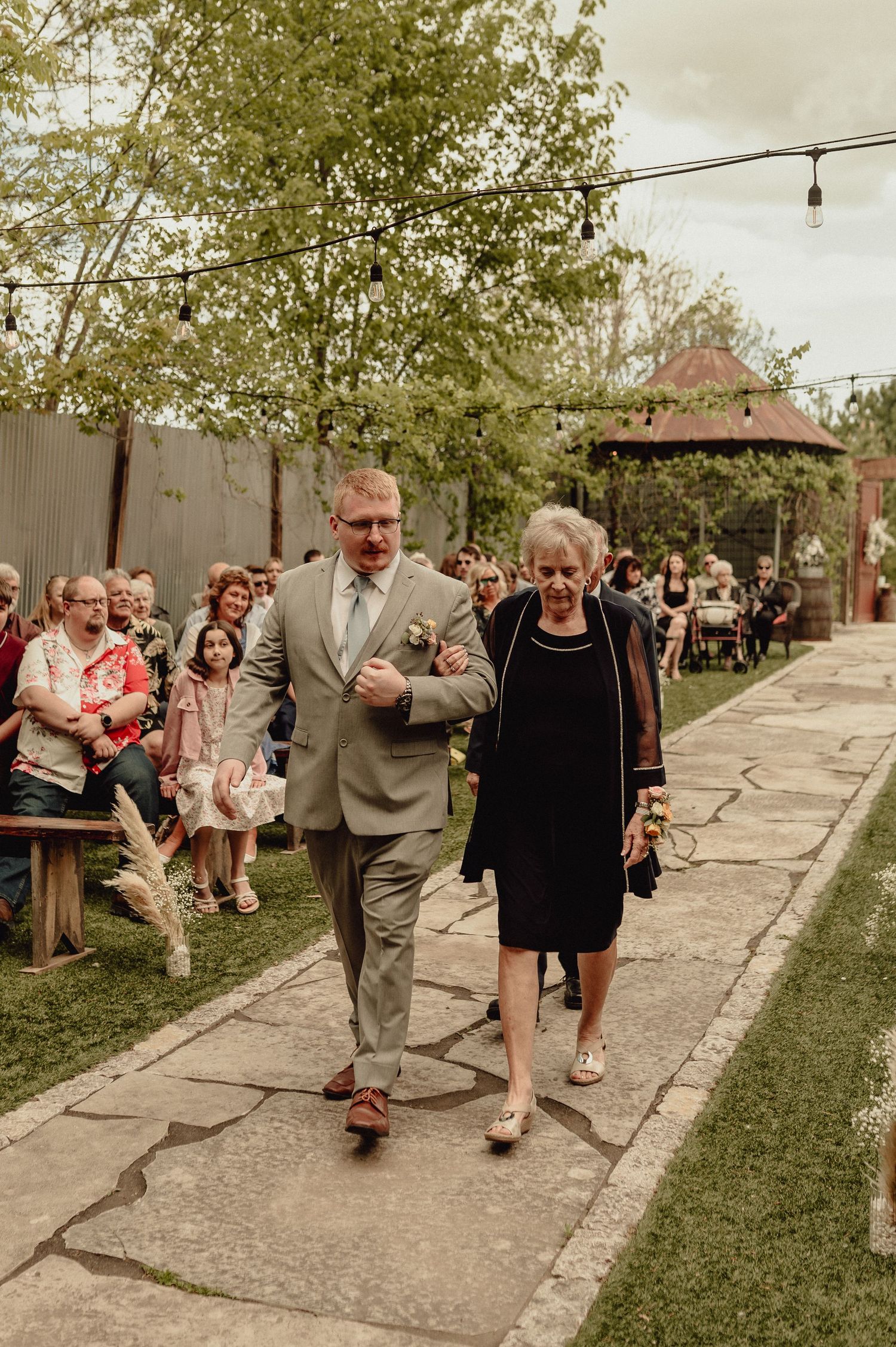 A couple walks down a stone path at an outdoor wedding ceremony while guests watch from wooden benches.