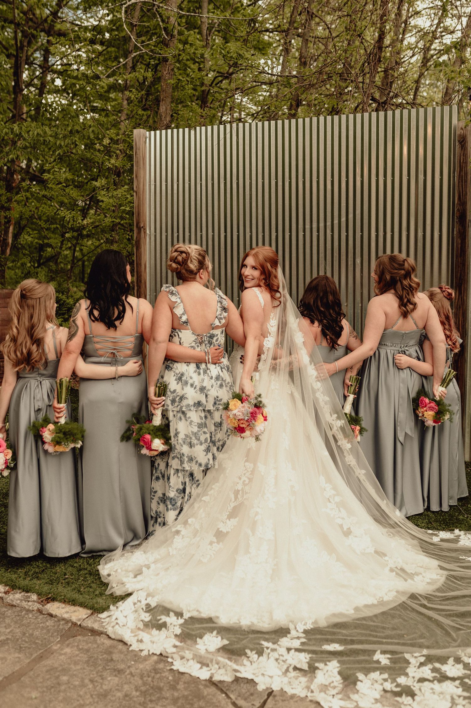 A wedding party in gray bridesmaid dresses and a white lace gown with a long train pose together outdoors.