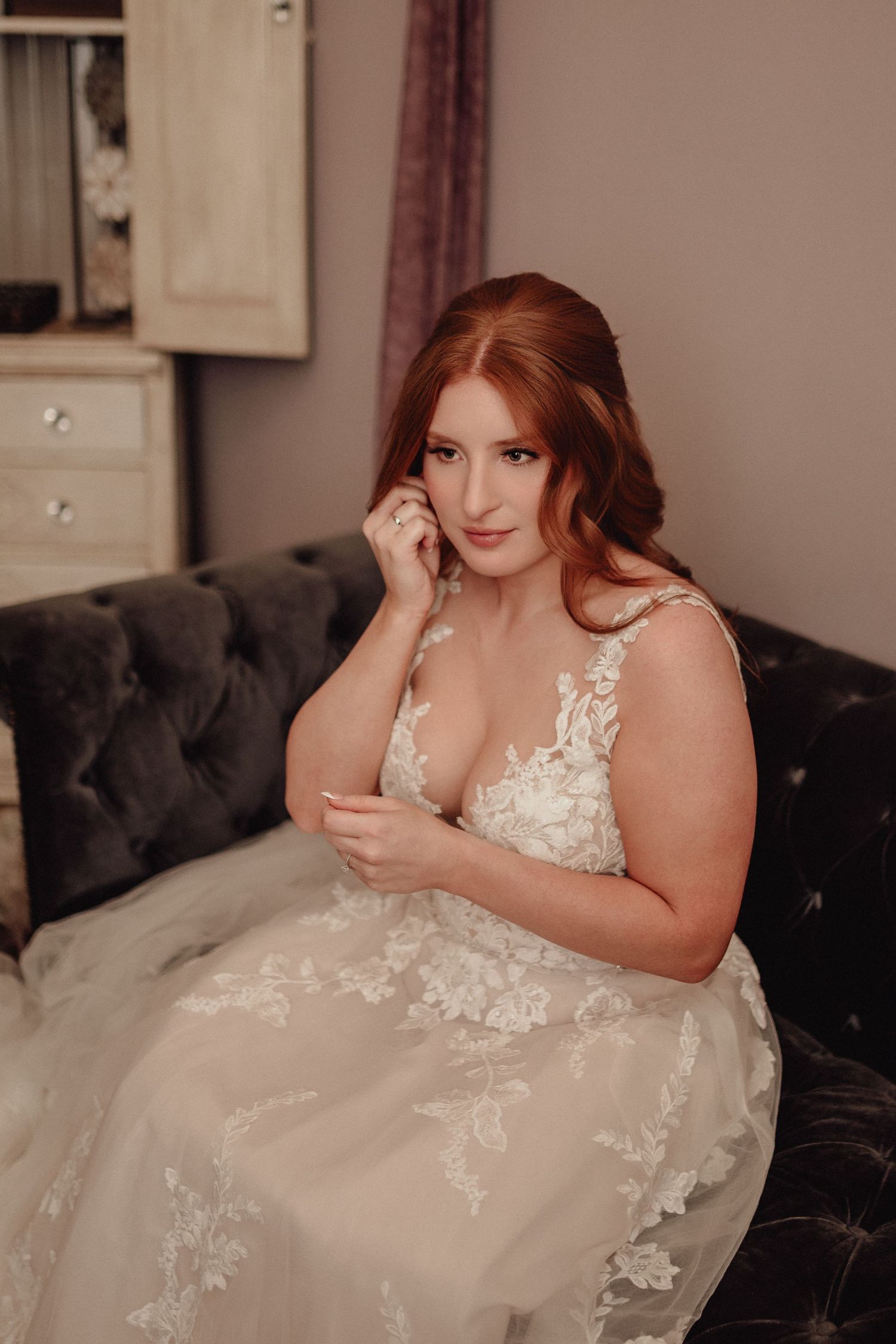 A bride in a white lace wedding dress with floral appliques poses elegantly on a dark velvet couch.